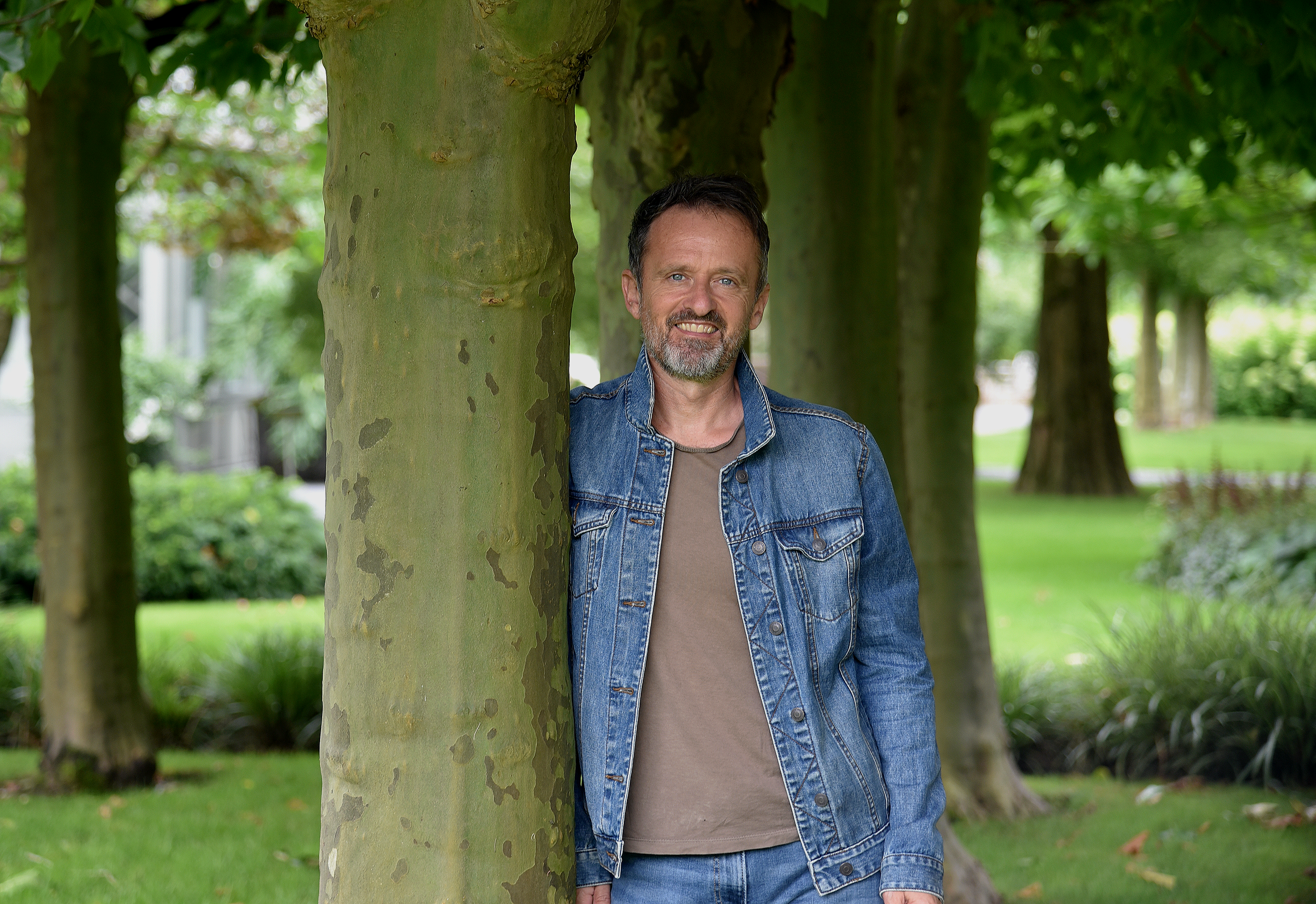 Mann lehnt an Baum im Park, lächelnd, umgeben von grünen Bäumen und Rasen.