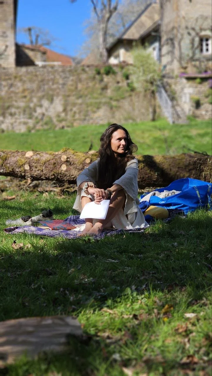 Frau sitzt im Grünen auf einer Decke, umgeben von Rucksack und Outdoor-Ausrüstung, mit einem Stück Papier in den Händen, im Hintergrund ein bewaldetes Gebiet und ein Haus, bei sonnigem Wetter.