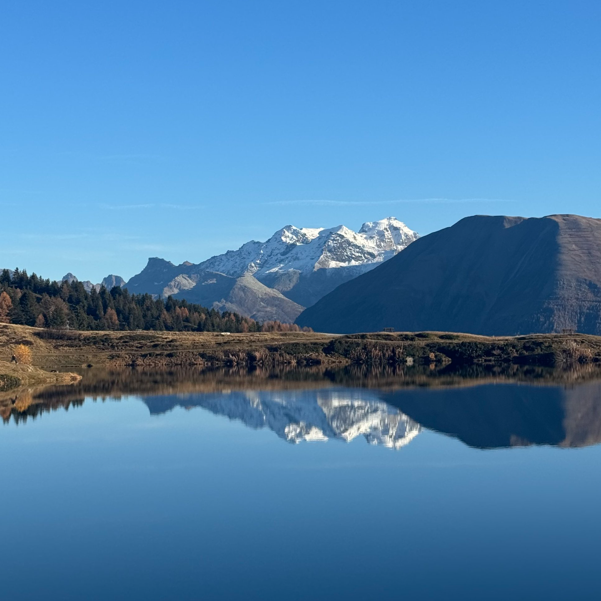 Berglandschaft mit schneebedeckten Gipfeln, Hügeln und einem See mit deutlich sichtbarer Spiegelung der Berge im Wasser bei klarem blauen Himmel.