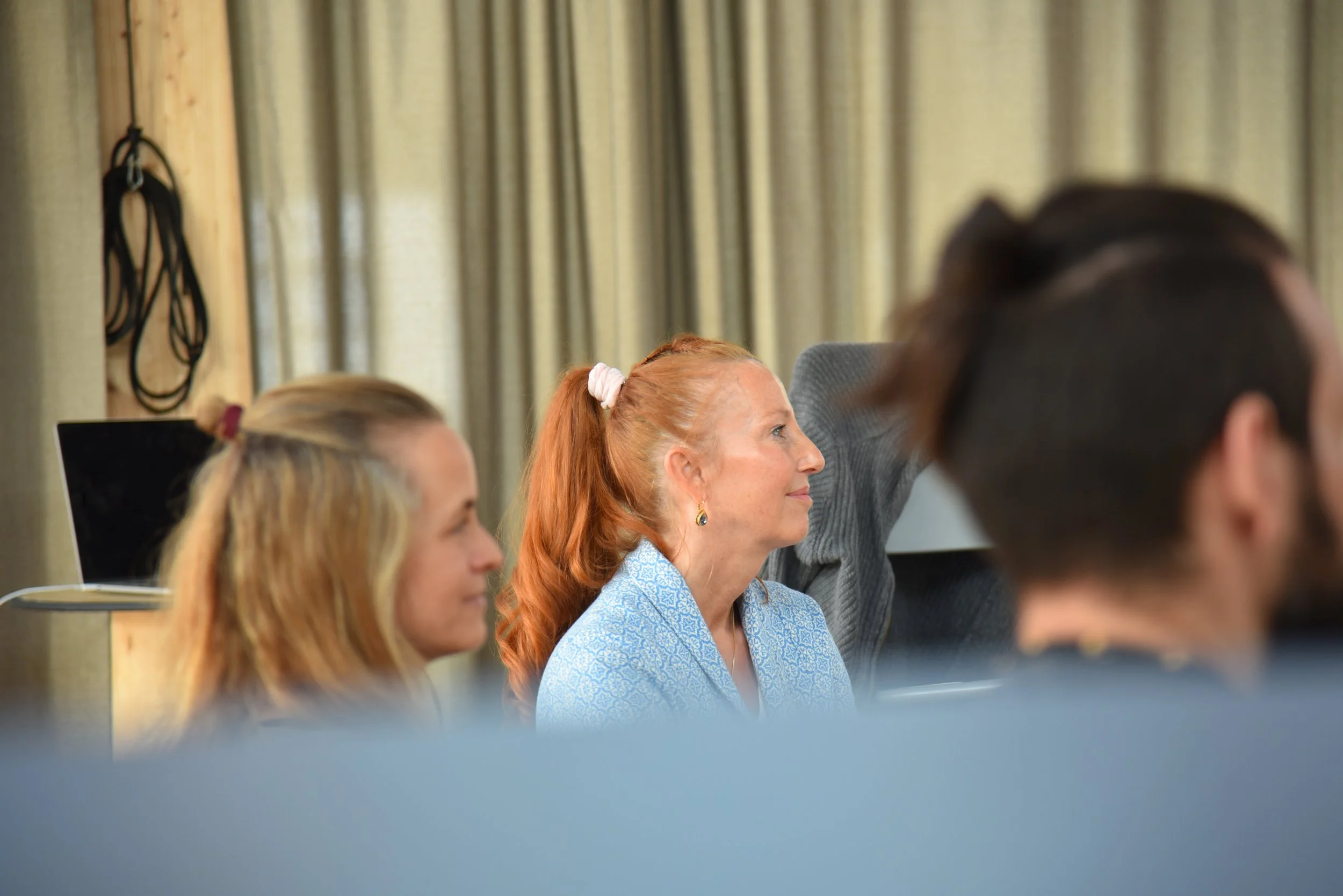 Mehrere Menschen sitzen in einem Raum bei einer Veranstaltung, fokus auf eine rot haarige Frau in blauer Jacke, die aufmerksam zuschaut.