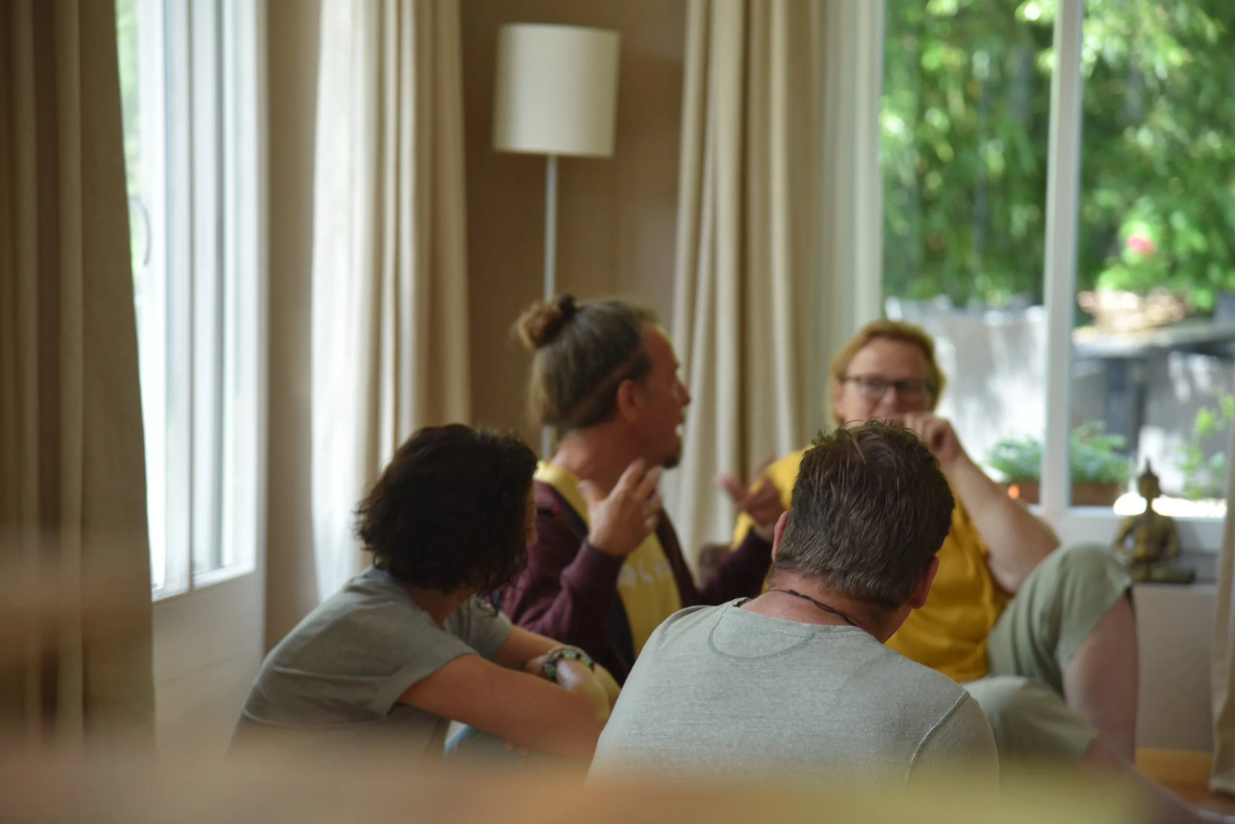 Sechs Menschen sitzen in einem hell erleuchteten Raum an einem Tisch, mit einem großen Fenster im Hintergrund, sprechen miteinander und diskutieren.