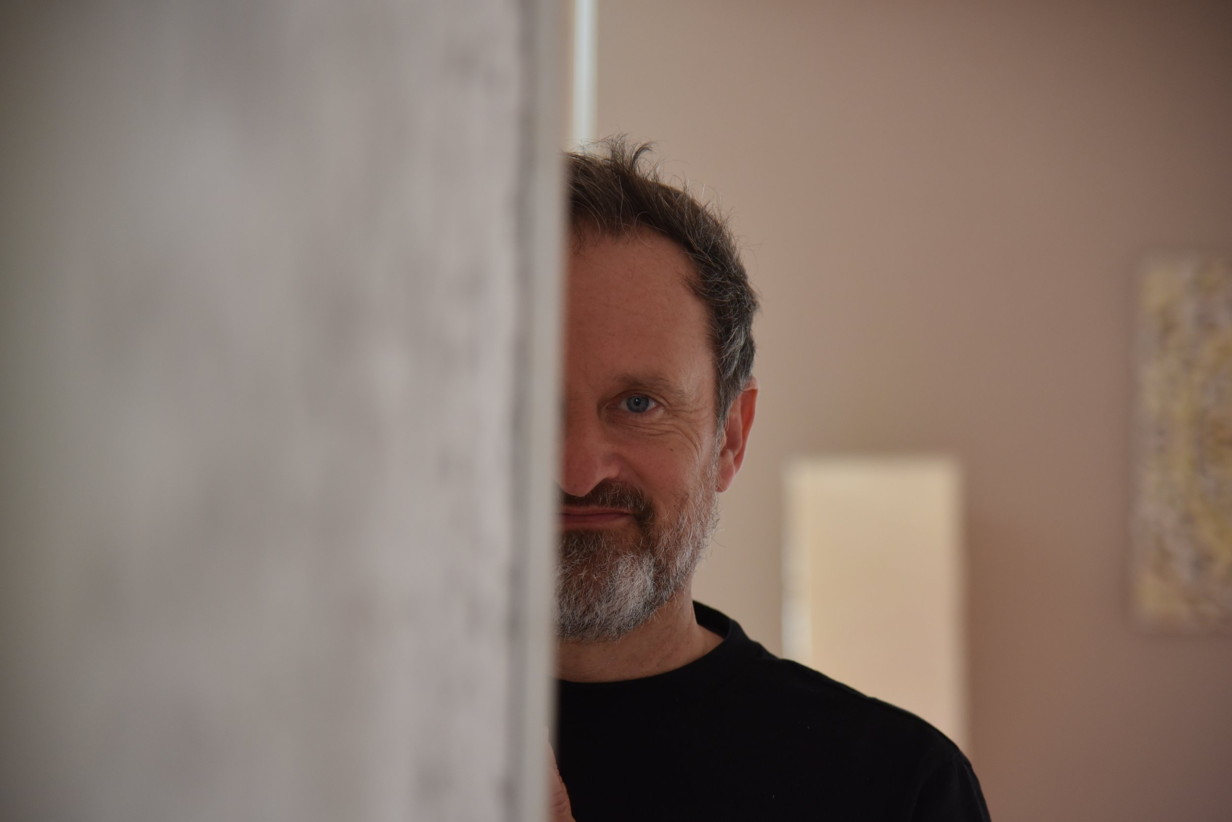 Ein Mann mit grauem Bart und braunen Haaren schaut neugierig um eine Ecke in einem schön beleuchteten Raum.