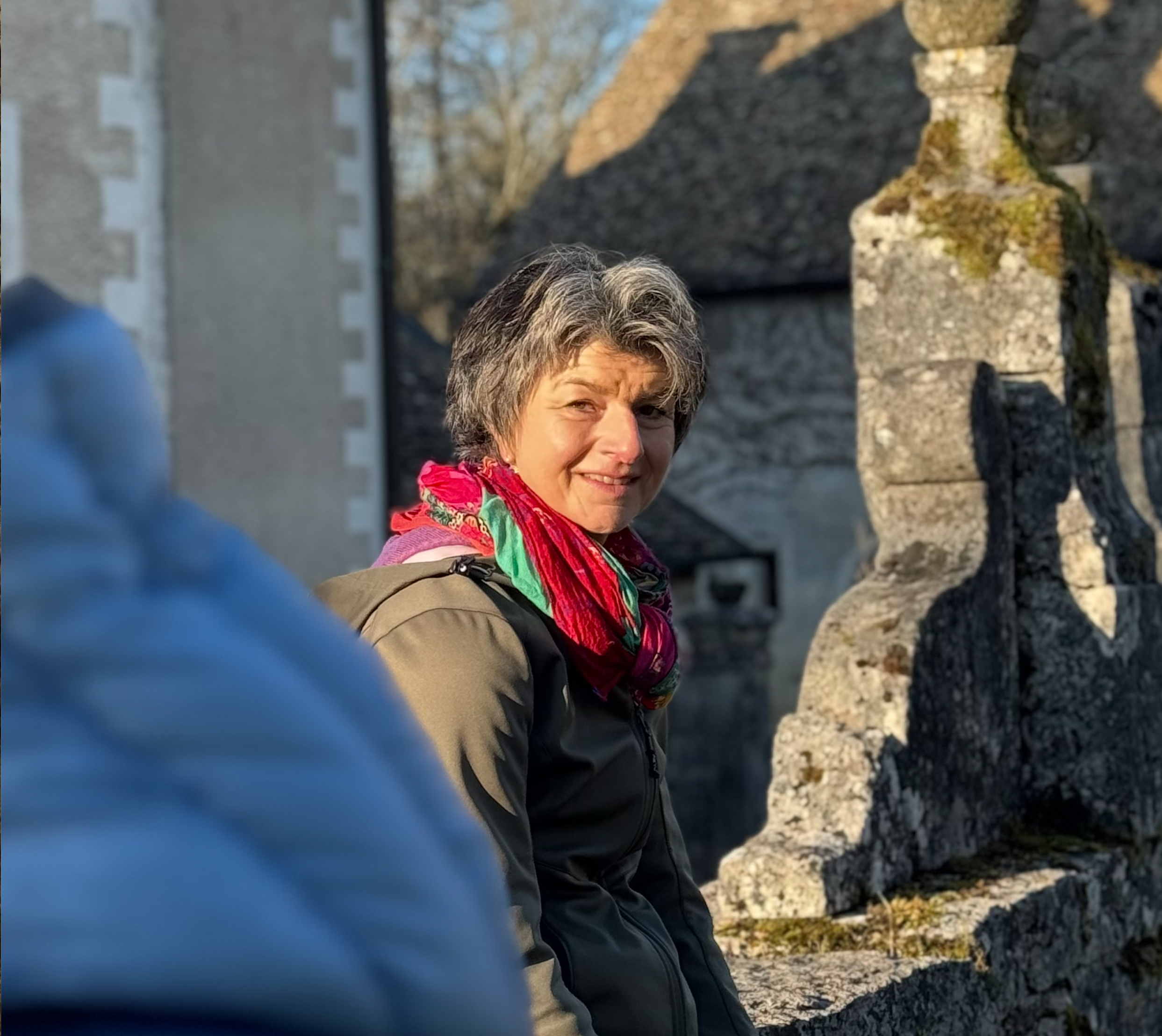 Eine ältere Frau mit grauem Haar, die draußen in der Sonne sitzt, trägt eine braune Jacke und einen roten, grünen und violetten Schal. Im Hintergrund sind alte Steinstrukturen und Bäume sichtbar.