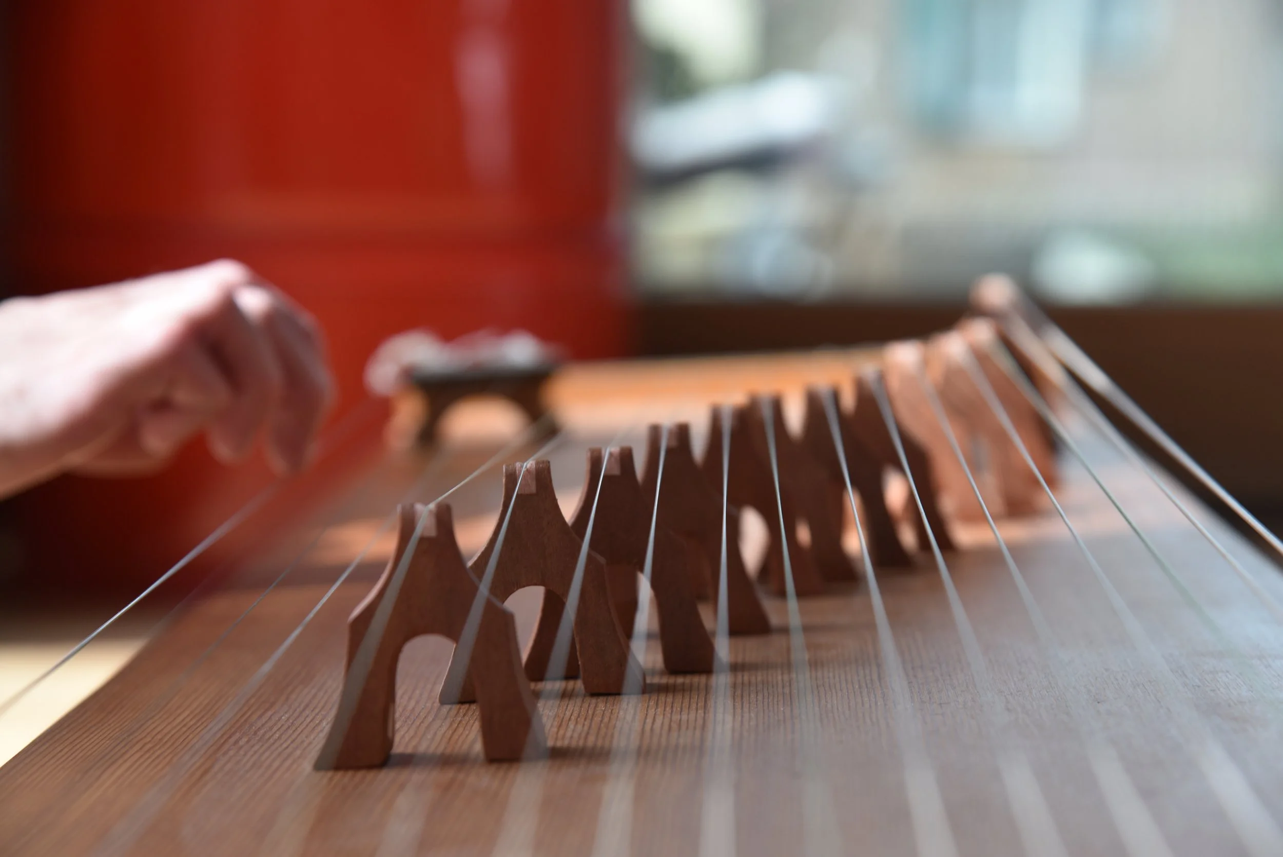 Nahaufnahme einer Person, die eine Zither spielt, mit Blick auf die Saiten und den Korpus aus Holz, im Hintergrund ein Fenster mit natürlichem Licht.