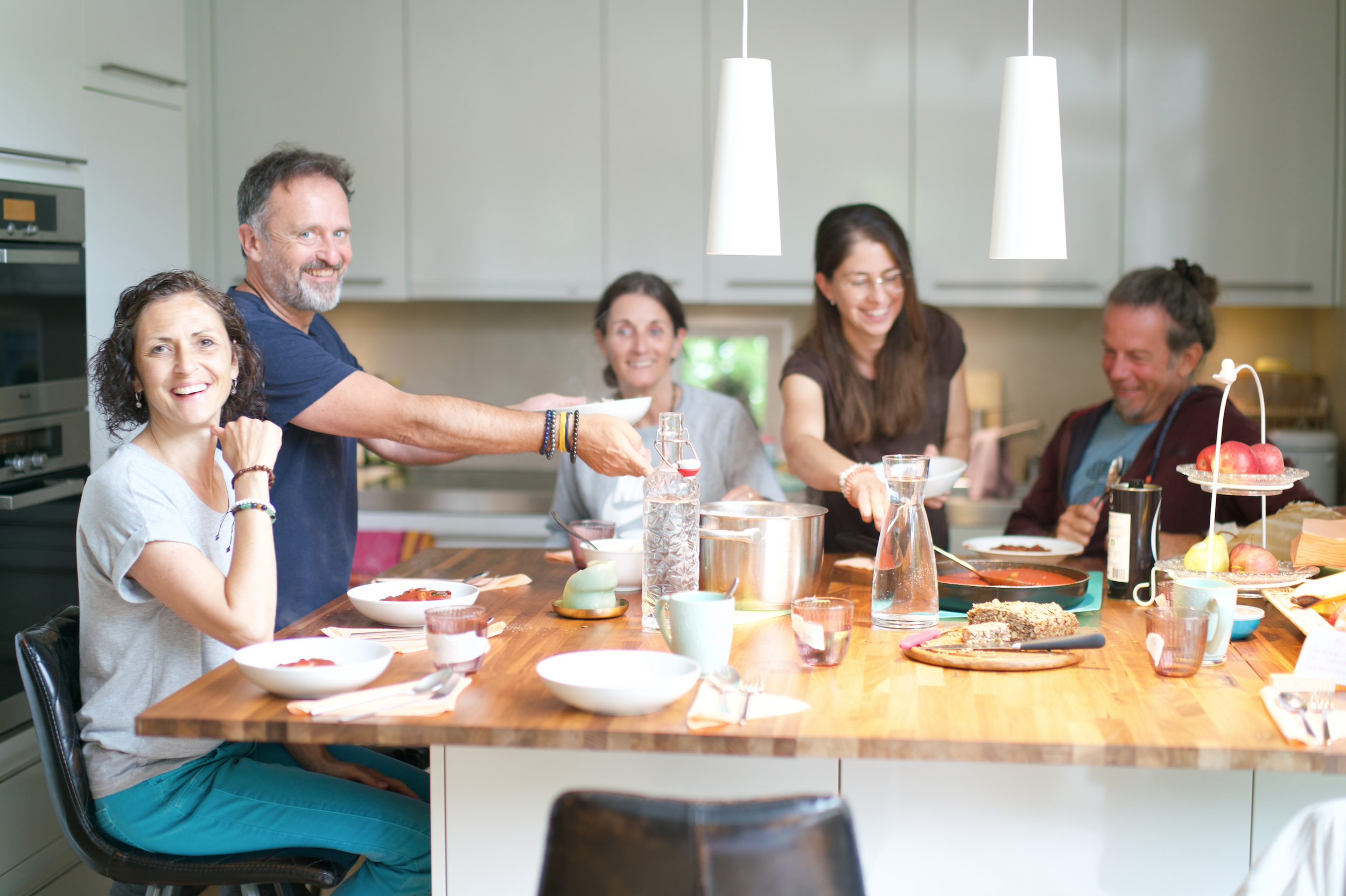Fünf Menschen beim gemeinsamen Essen in einer modernen Küche, lachen und teilen Speisen, auf dem Tisch sind Teller, Gläser und Gebäck.
