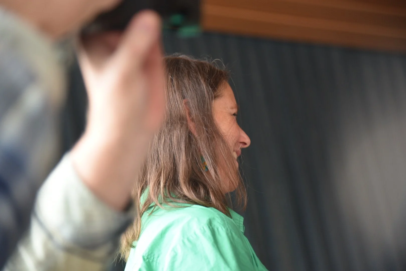 Frau mit braunen, lockigen Haaren, lachend, in einem hellgrünen Hemd, im Hintergrund ein dunkler Vorhang.