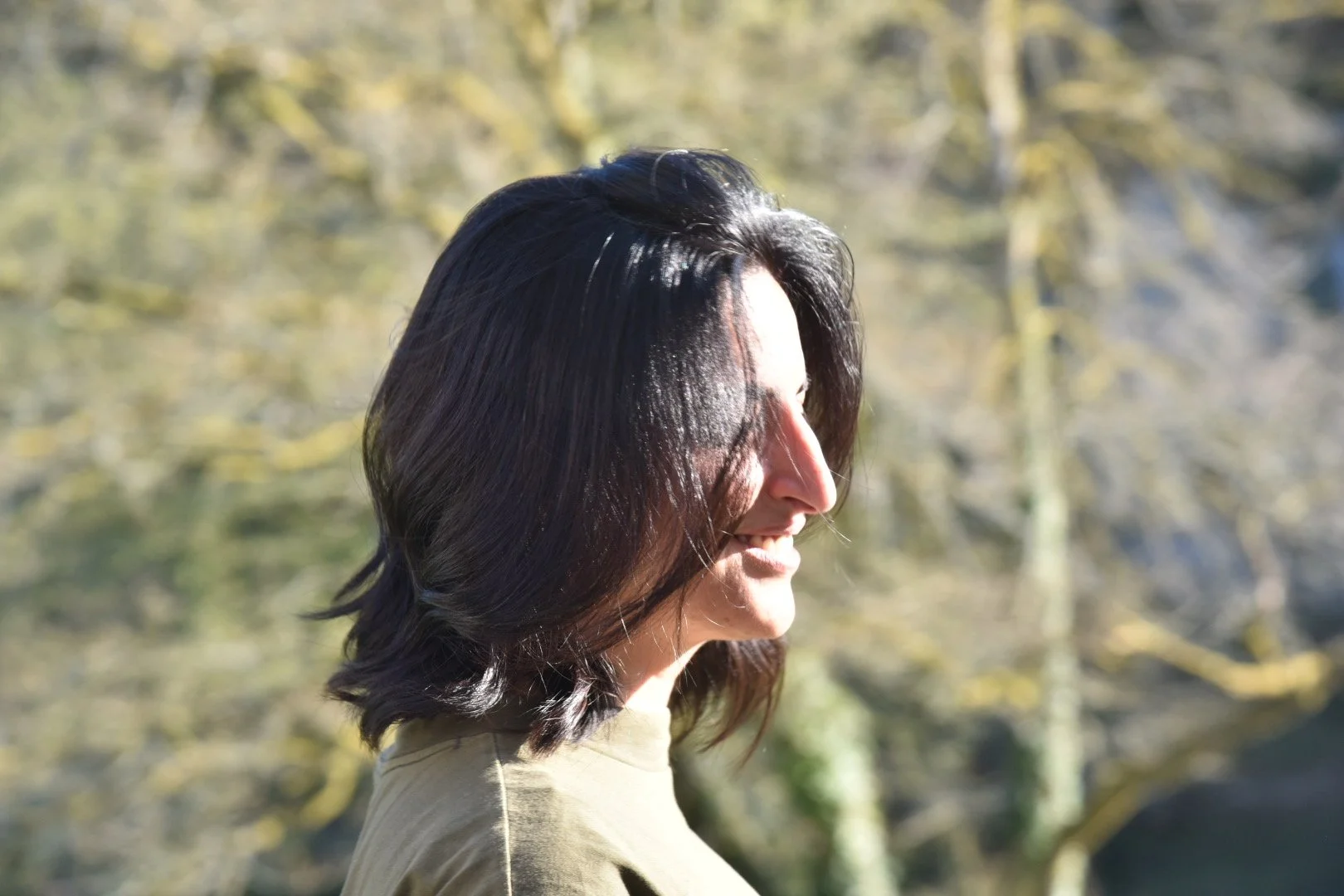 Seitenansicht einer lächelnden Frau mit schwarzen, welligen Haaren im Freien bei Sonnenlicht, im Hintergrund Bäume.