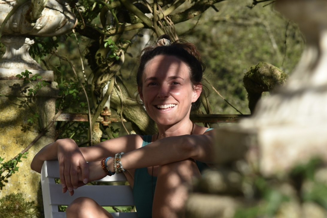 Eine lachende Frau sitzt in einem Garten, umgeben von Pflanzen und alten Steinskulpturen, mit Sonnenlicht, das Schatten wirft.