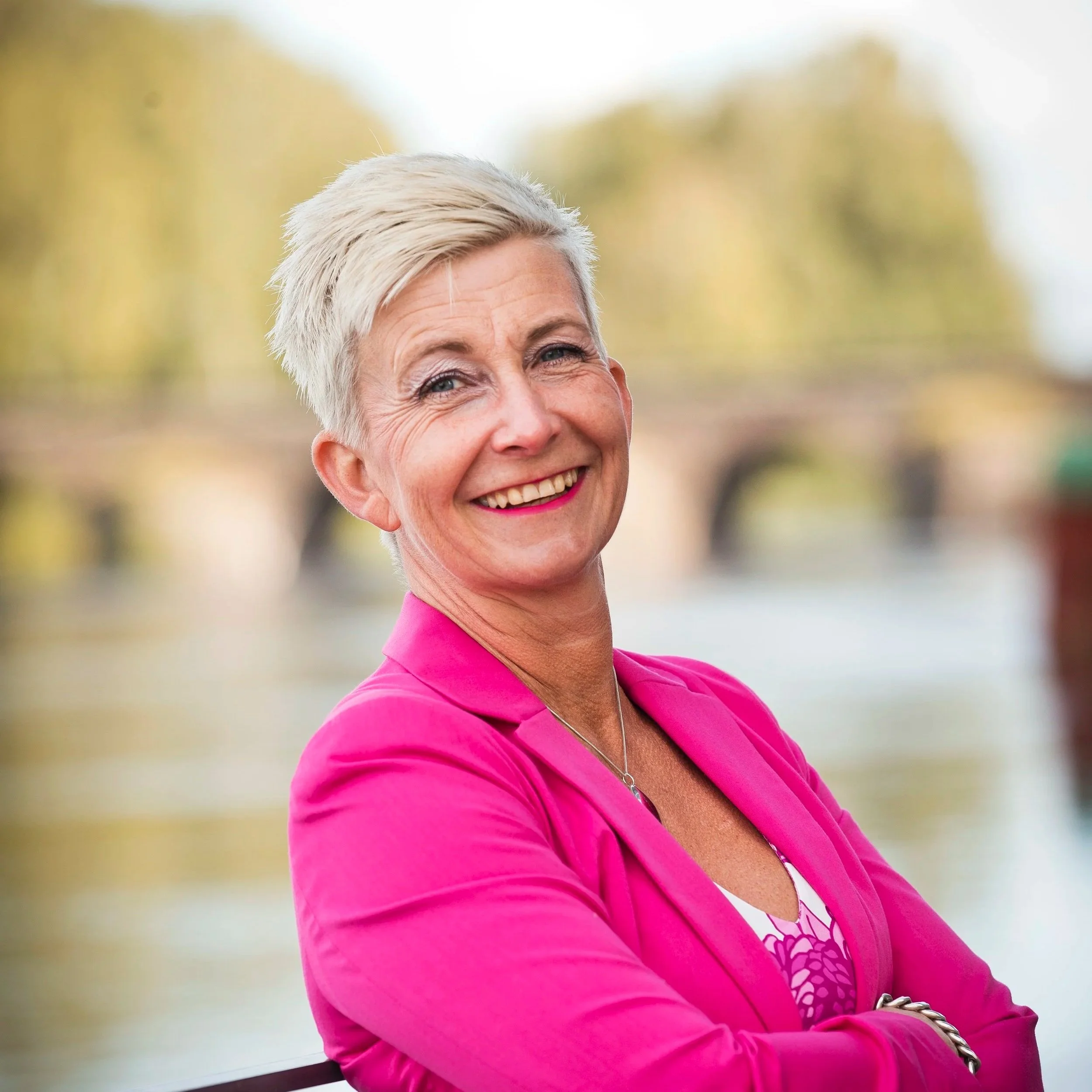 En leende kvinna med kort, ljusblond hår, bär en rosa blazer, står framför en vacker, lugn vattenmiljö med broar och gröna träd i bakgrunden.