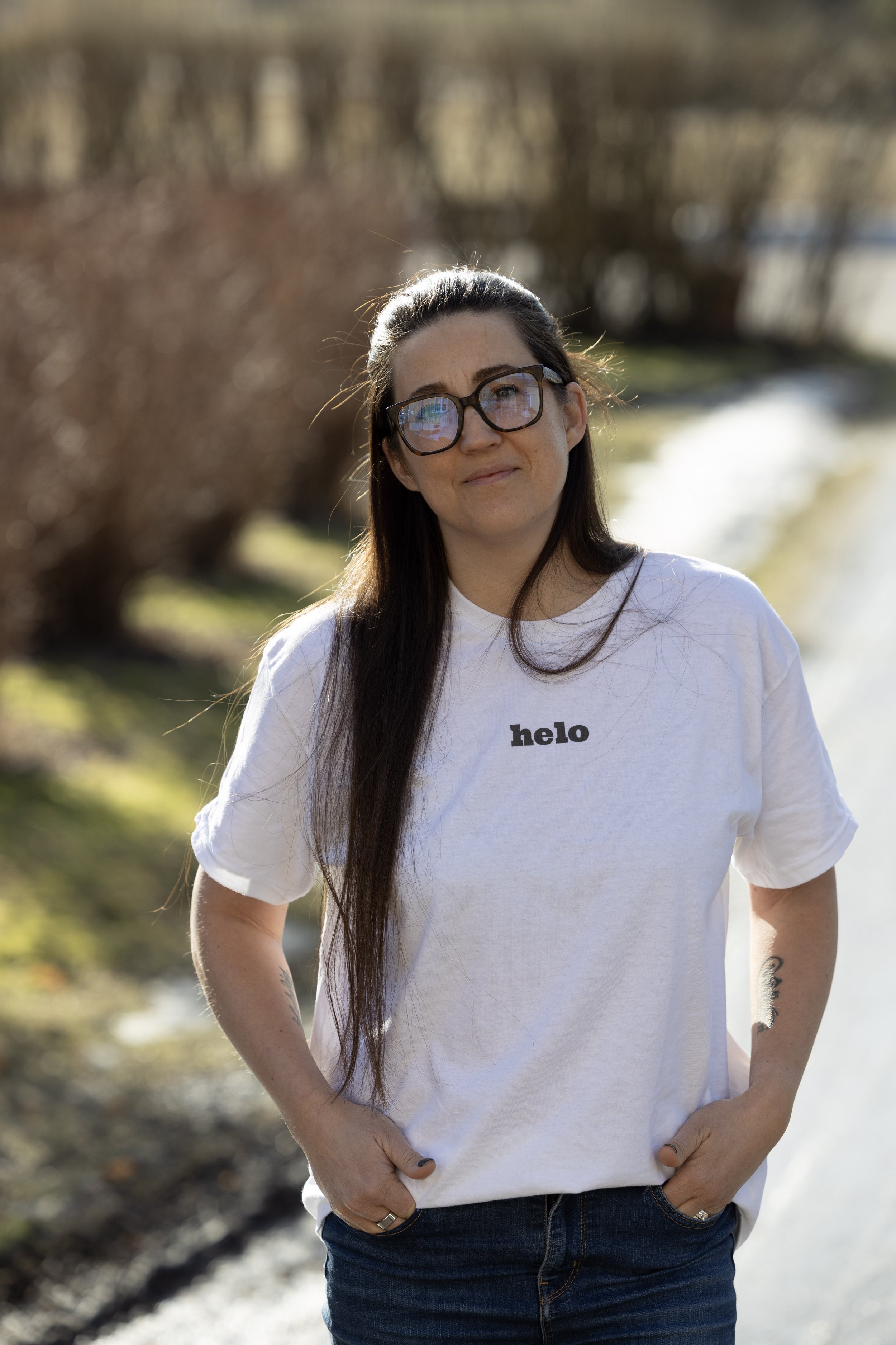 En kvinna står utomhus i solljus, bär glasögon och en vit t-shirt med texten 'helo'.