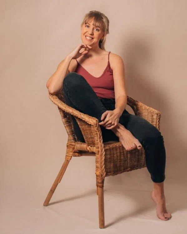 Ung kvinna sitter i en rottingstol, ler och har benet korsat över det andra, klädd i en rosa topp och svarta jeans.