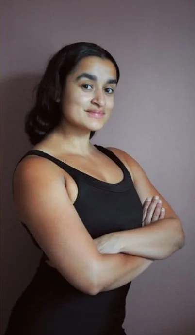 Kvinna i svart ärmlös topp som står med armar korsade, mot en lila vägg.