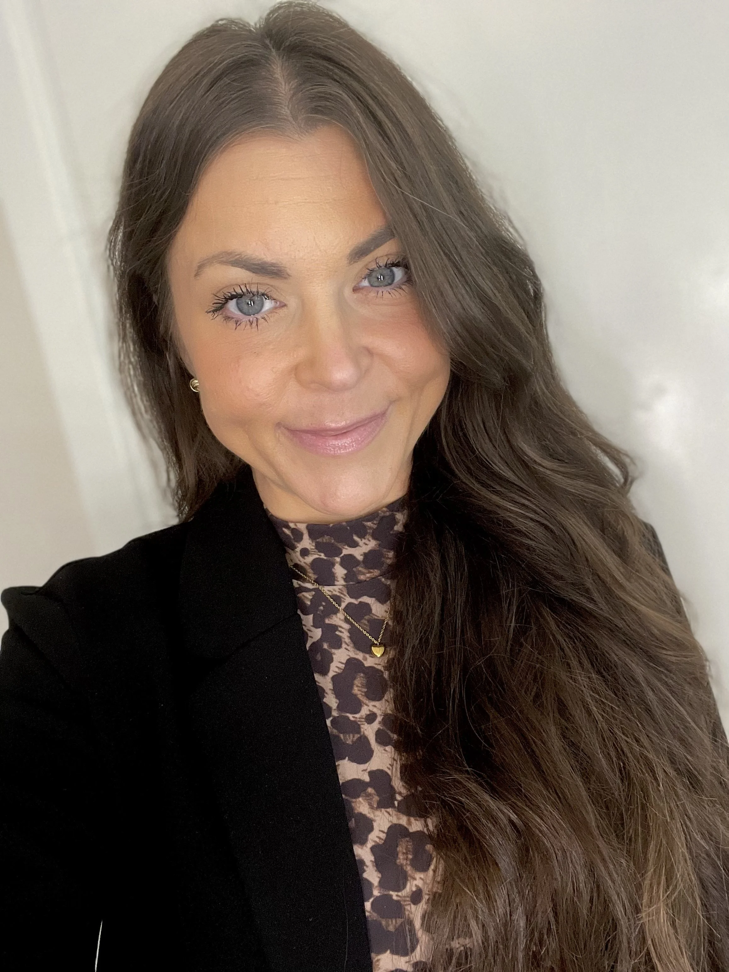 En kvinna med långt, lockigt brunt hår, ljus hud och blå ögon, klädd i en leopardmönstrad top och svart blazer, tar en selfie framför en vit vägg.