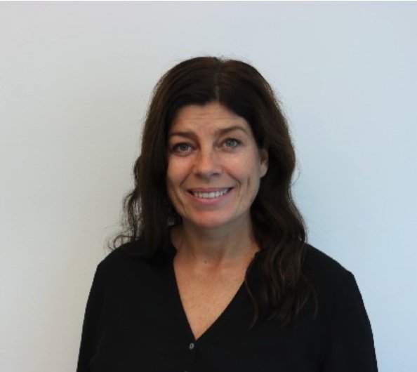 En kvinna med mörkt, vågigt hår och blå ögon, som ler och bär en svart tröja, står framför en vit vägg.