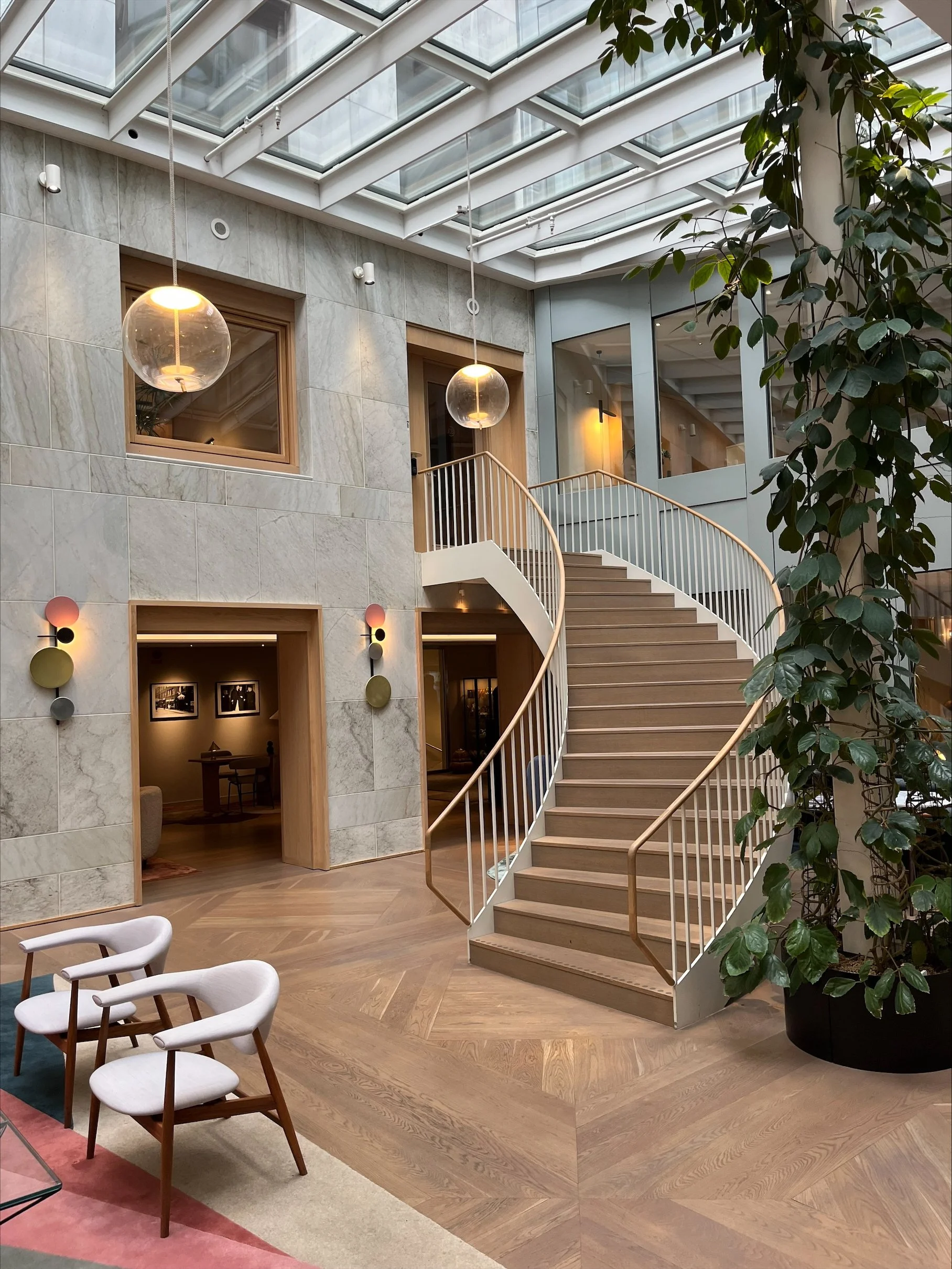 Inomhusmiljö med en spiraltrappa i trä, stora fönster i taket, växter och samtalsstolar.