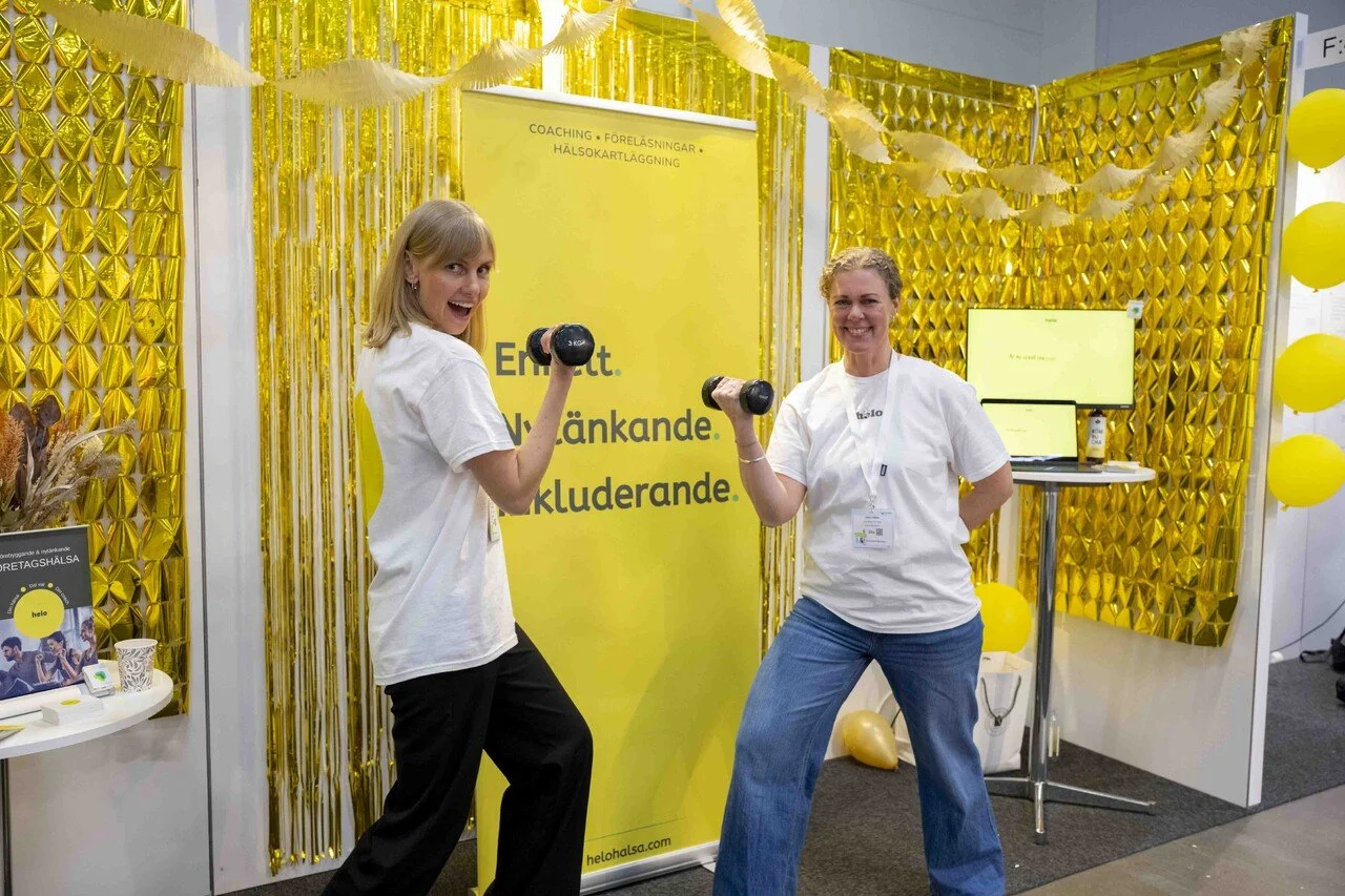 Två kvinnor i vita tröjor och jeans som tränar med hantlar framför en guldklädd vägg på ett mässställ. Bakom finns en gul skylt med text och en skärm.