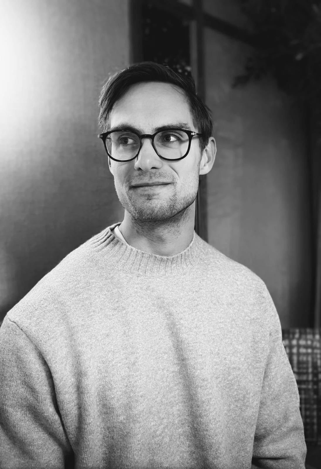 En ung man med glasögon, mörkt hår och en ljus tröja, tittar åt sidan i svartvitt.
