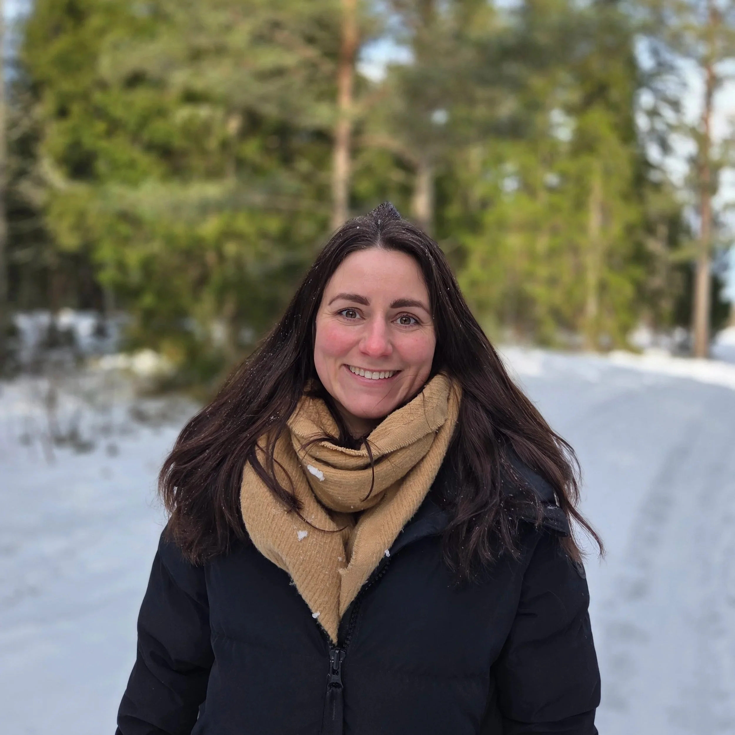 En kvinna med brunt hår och skinnjacka står i snön med träd och snöiga landskap i bakgrunden, ler och tittar mot kameran.