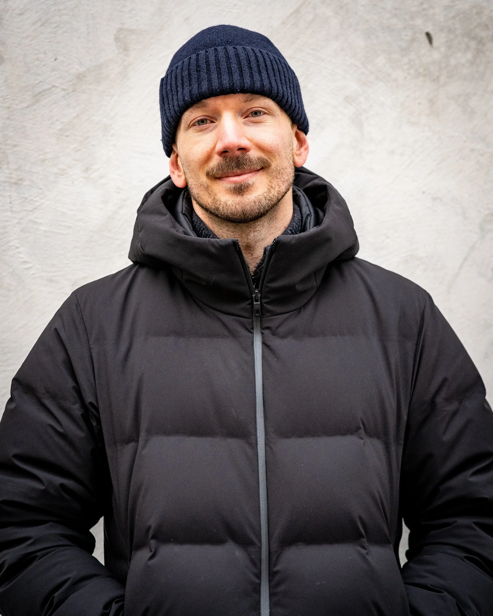 Man i svart jacka och blå mössa står mot en ljus vägg.