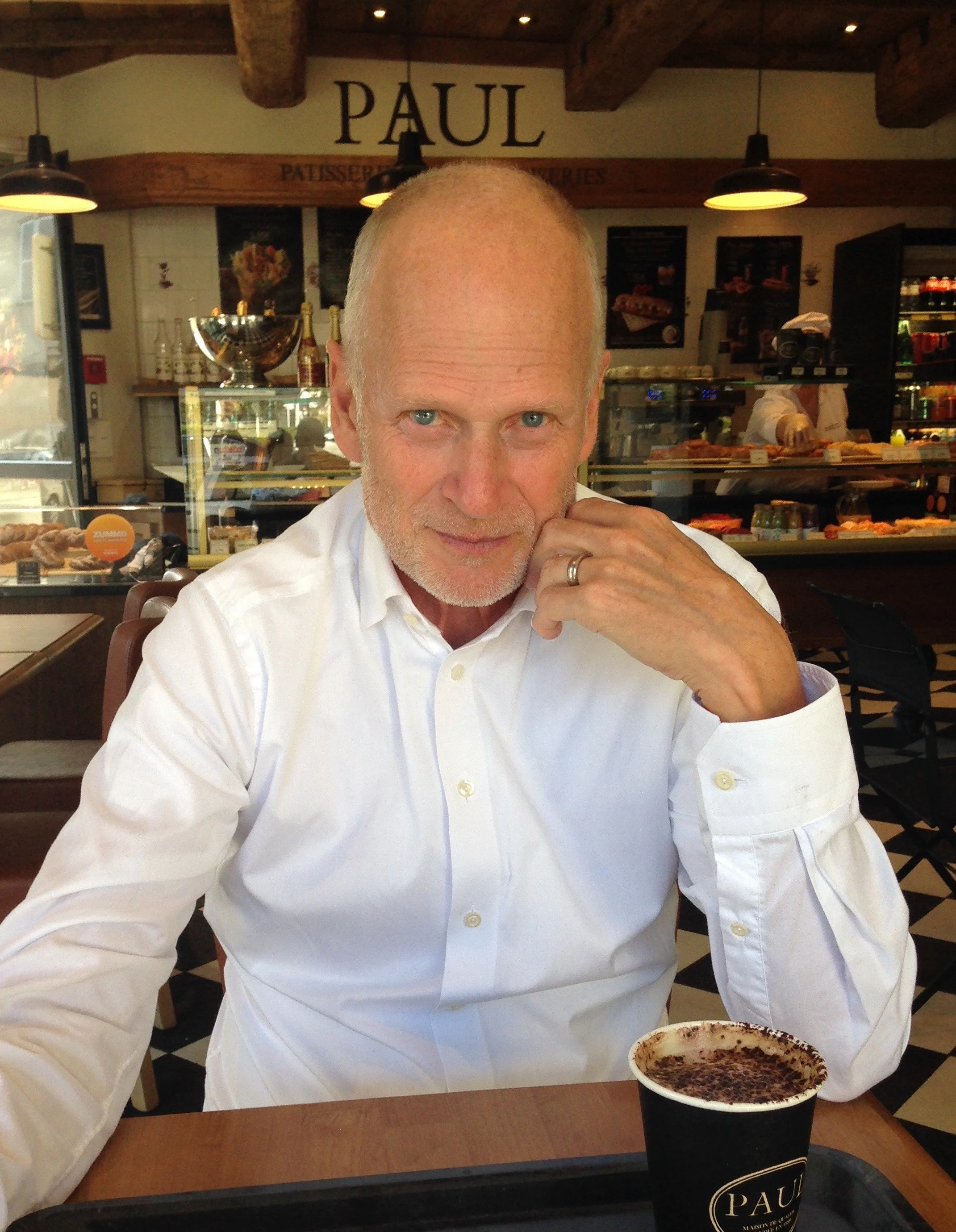 En äldre man i vit skjorta som sitter vid ett bord i ett café eller bageri, med en kopp varm choklad framför sig. Bakom honom syns en glasdisk med bakverk och ett skylt som säger 'PAUL'.