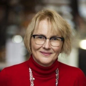 En kvinna med blont hår och glasögon, bär en röd tröja och ett pärlhalsband, står utomhus.