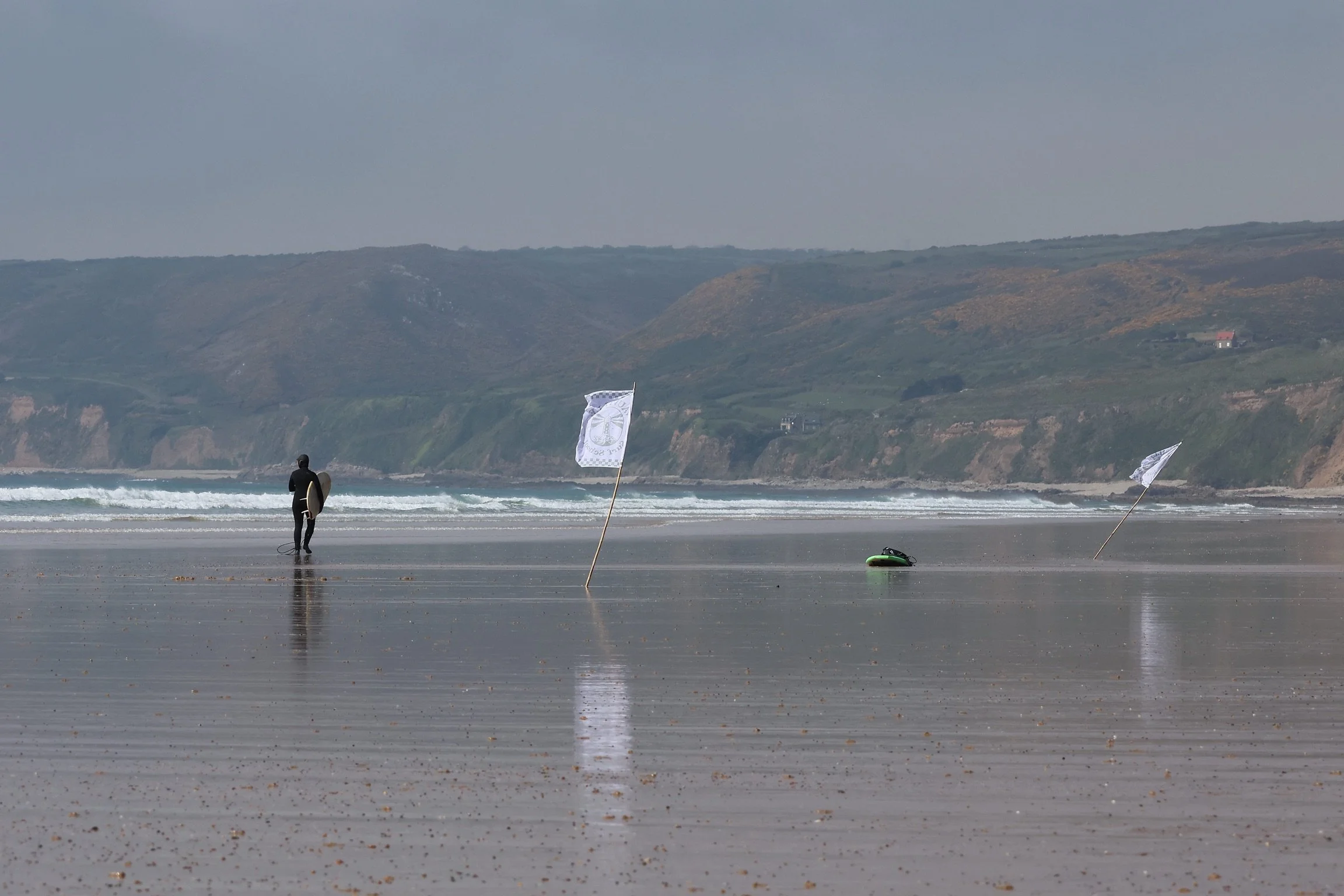 Une personne en combinaison avec une planche de surf à la main se trouve sur la plage. Il y a des drapeaux blancs plantés dans le sable et l'océan en arrière-plan, avec des collines verdoyantes au loin.
