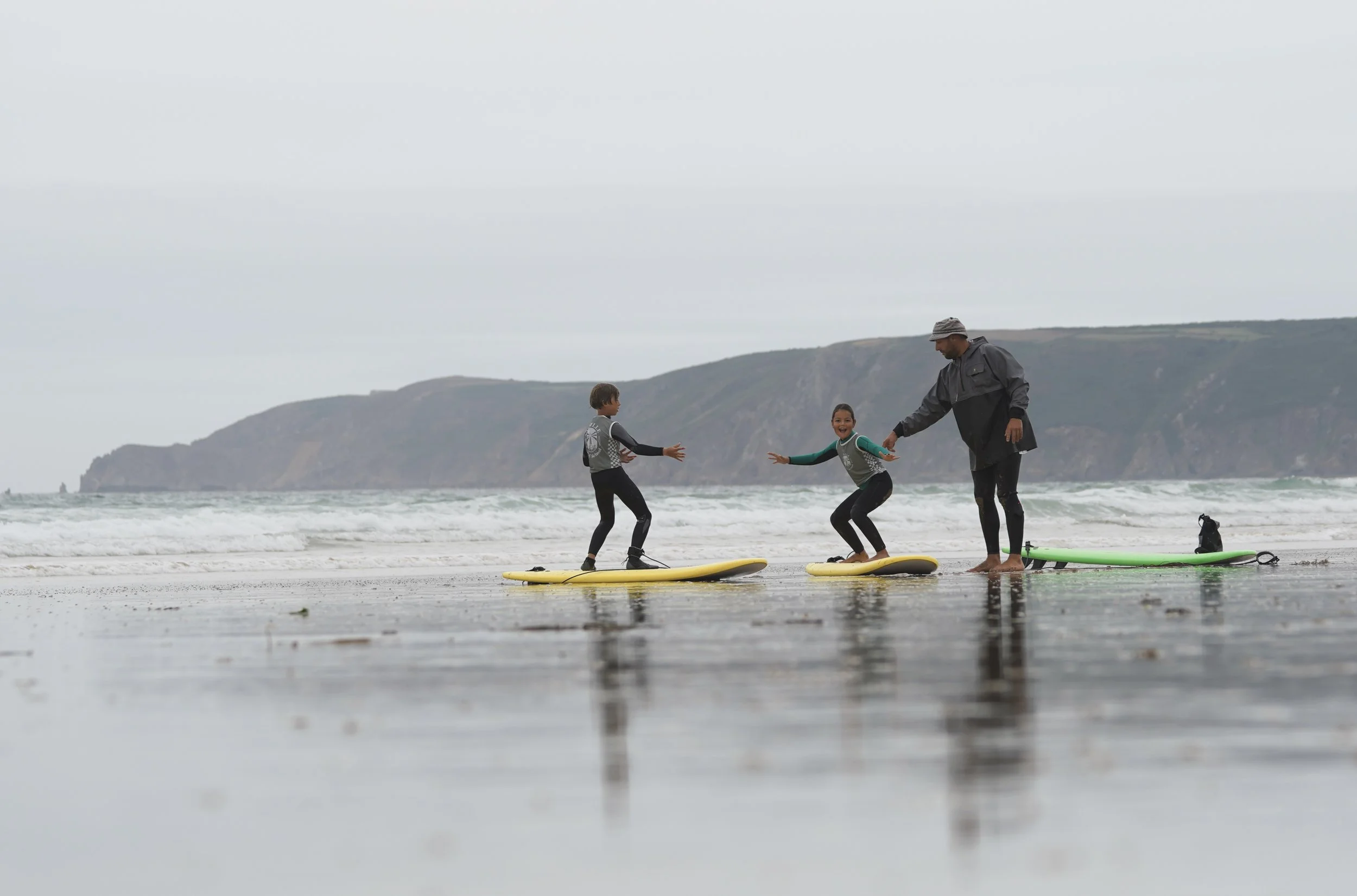 Un adulte et deux enfants sur la plage, en train de faire du surf, avec le cap de la Hague en arrière-plan, par temps nuageux.