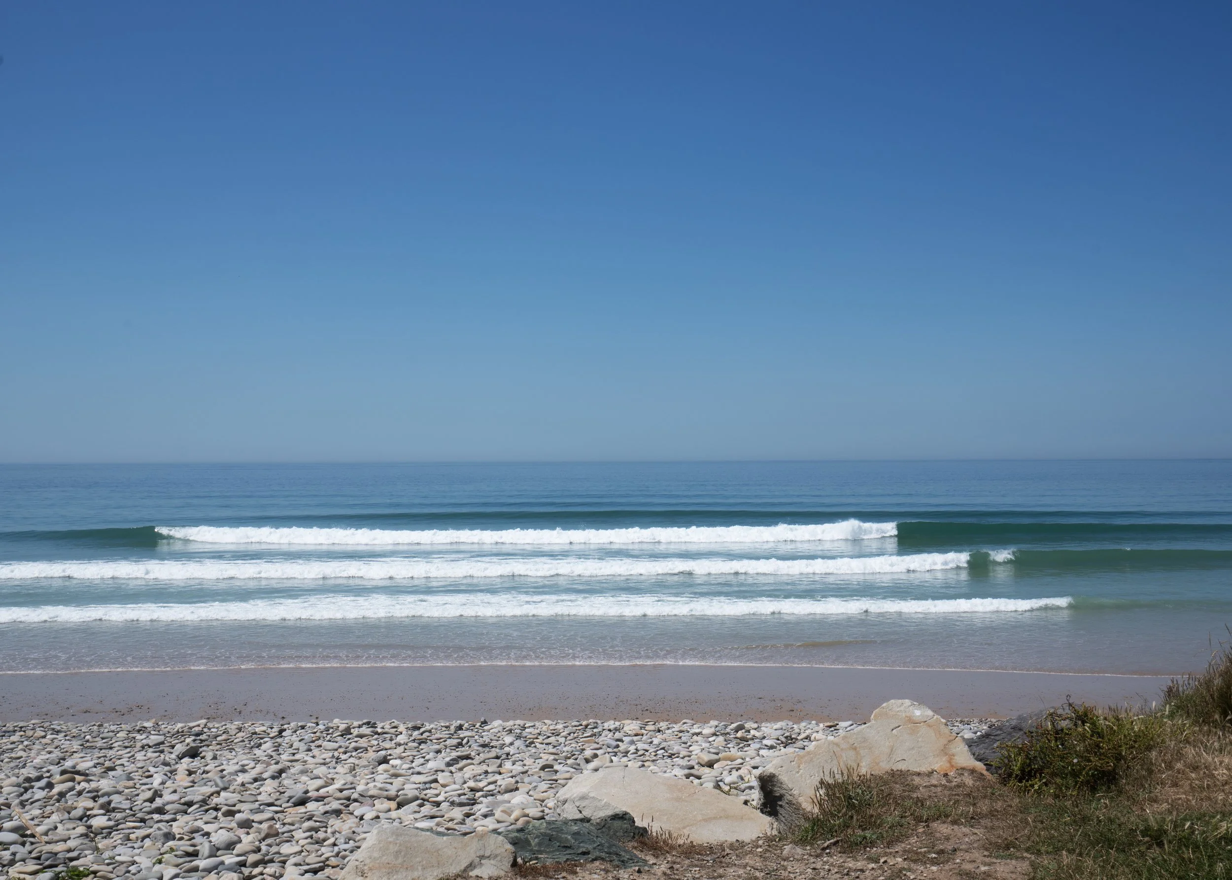 Plage avec sable, petits galets, plusieurs très belles vagues, ciel clair et horizon visible.