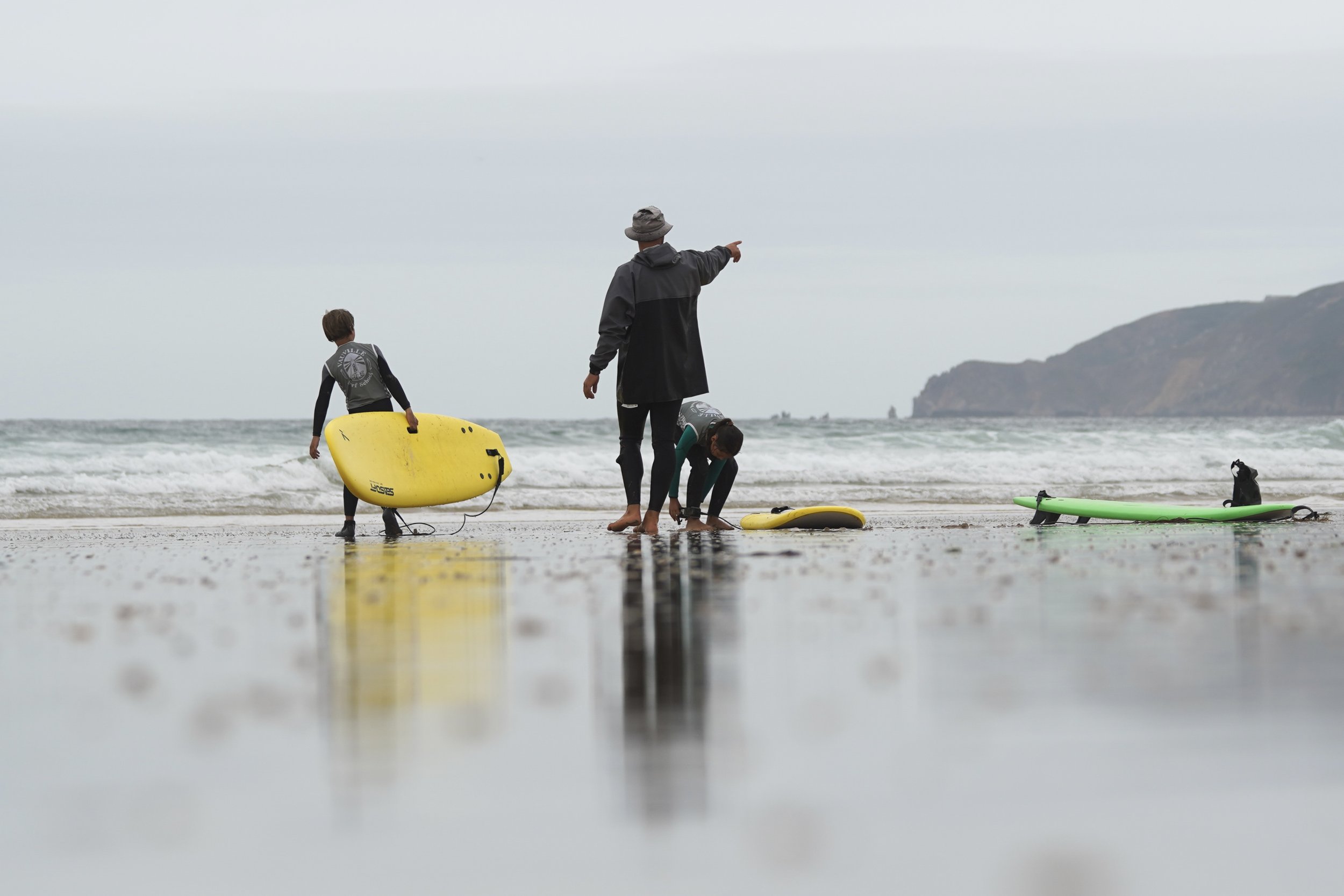Un adulte et deux enfants sur la plage, en train de préparer ou de ranger des planches de surf, sous un ciel nuageux.