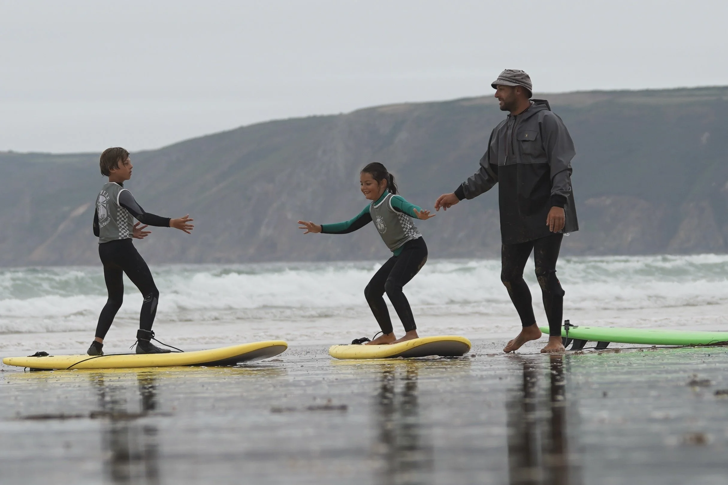 Un adulte et deux enfants en train de faire du surf sur la plage, avec le cap de la Hague en arrière-plan, sous un ciel nuageux.