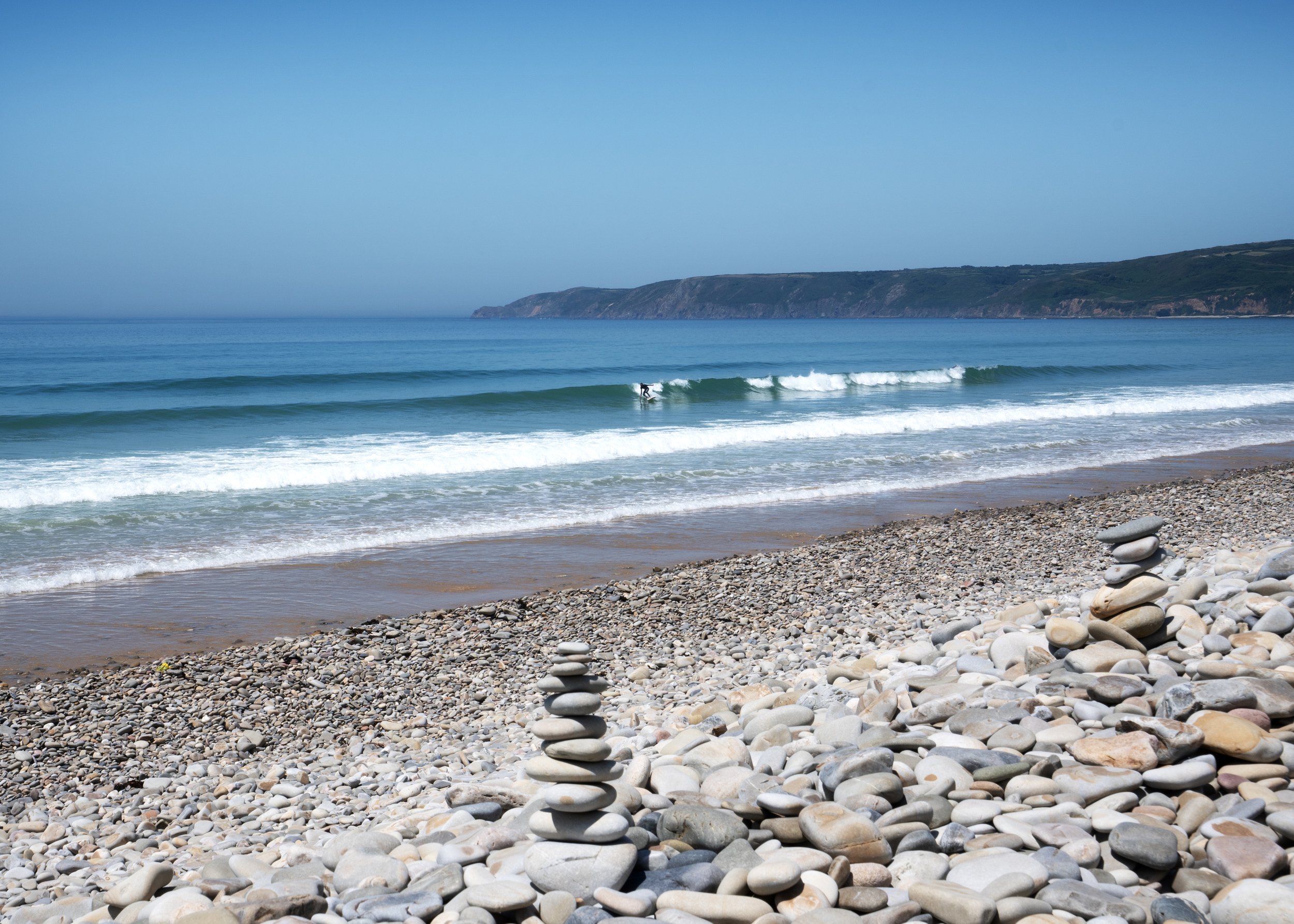 Plage avec des galets, un surfer sur une vague, et une côte rocheuse en arrière-plan sous un ciel clair.