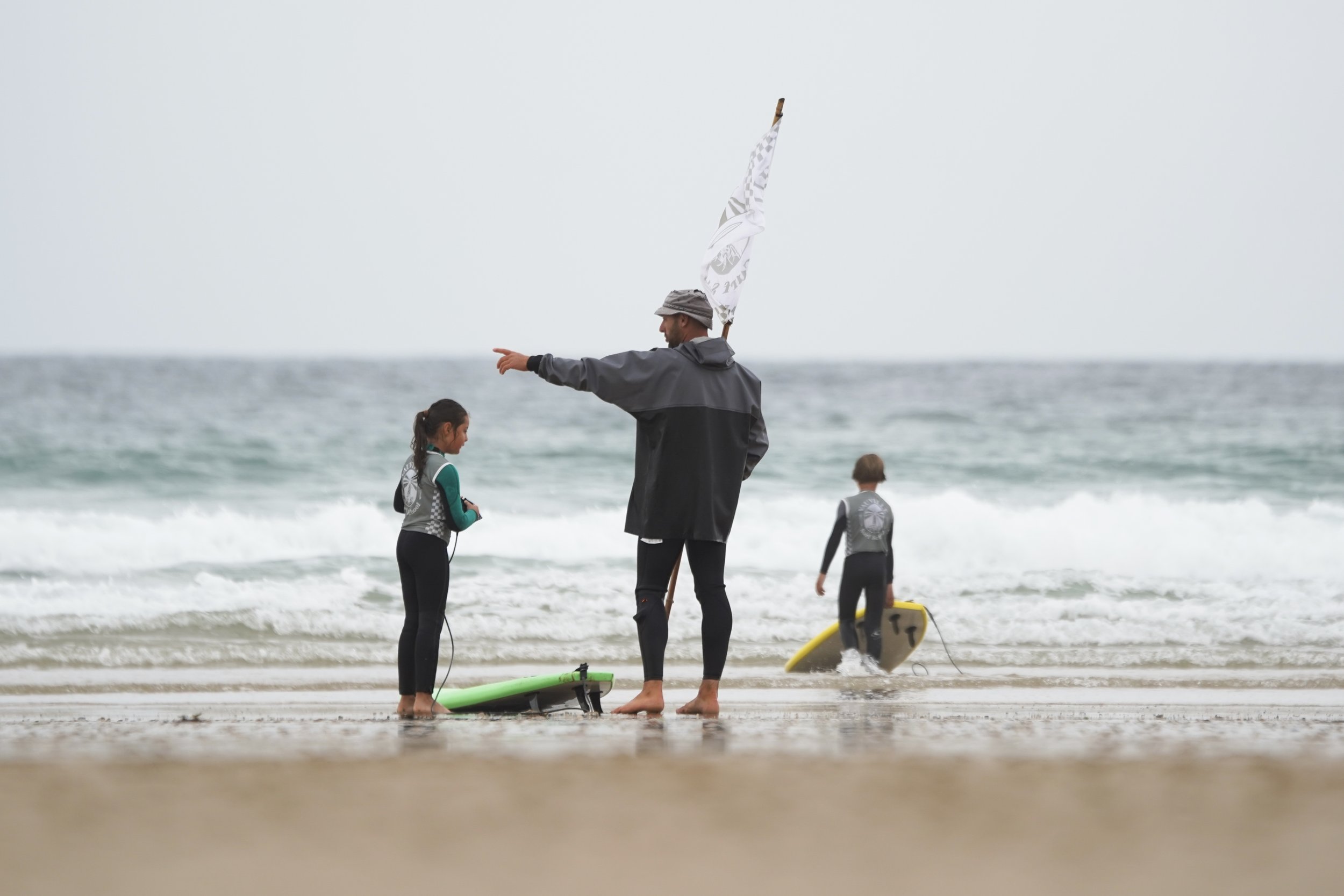 Personne enseignant à deux enfants comment faire du surf sur la plage, avec d'autres enfants en arrière-plan, près de l'eau et des planches de surf.