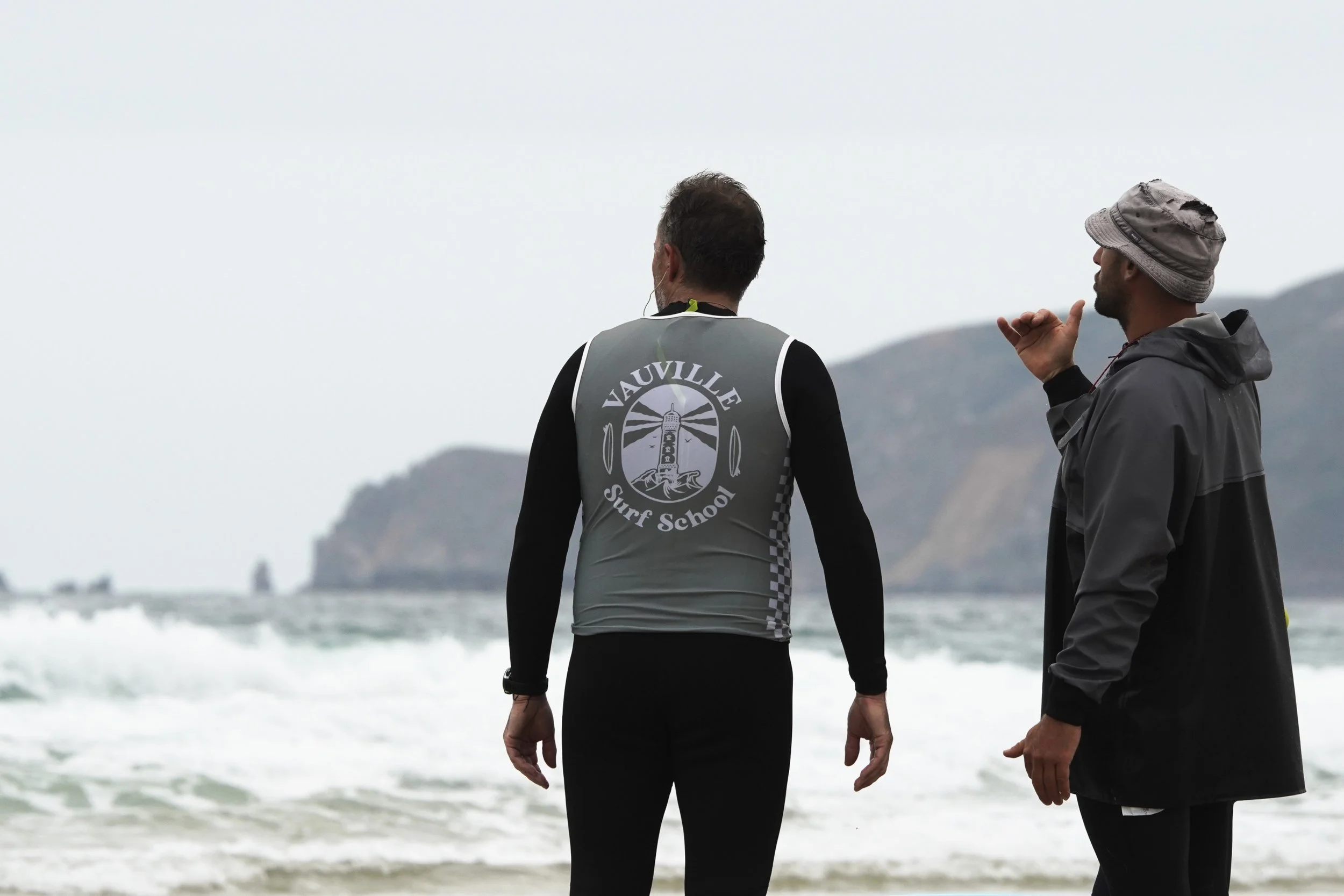Deux hommes, vus de dos, parlent sur la plage, avec la mer et des montagnes en arrière-plan, un homme porte un gilet avec le logo 'Vauville Surf School'.