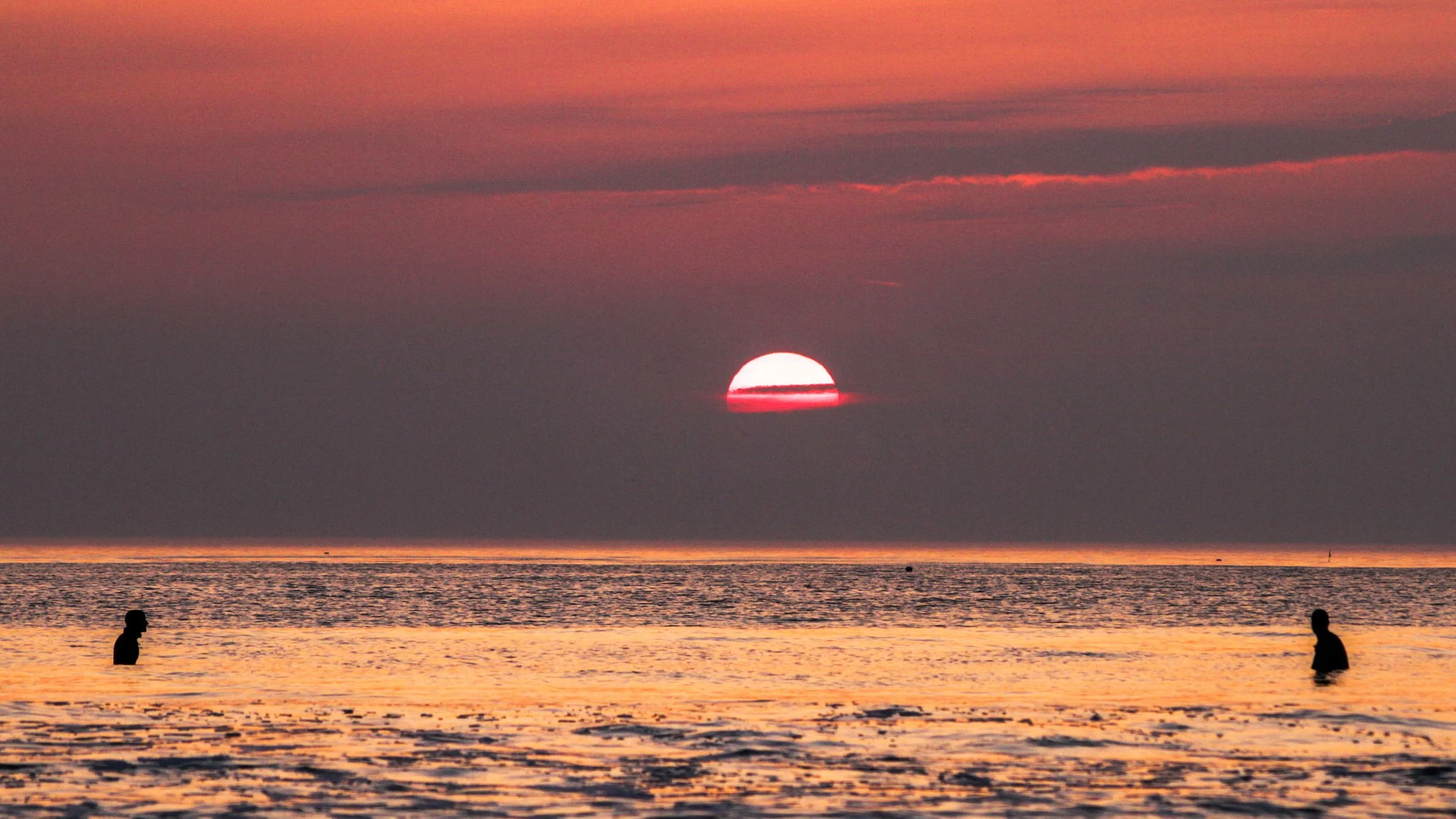 Coucher de soleil sur l'océan avec deux personnes attendant une vague assis sur leurs planche, ciel avec nuances d'orange et de rose.