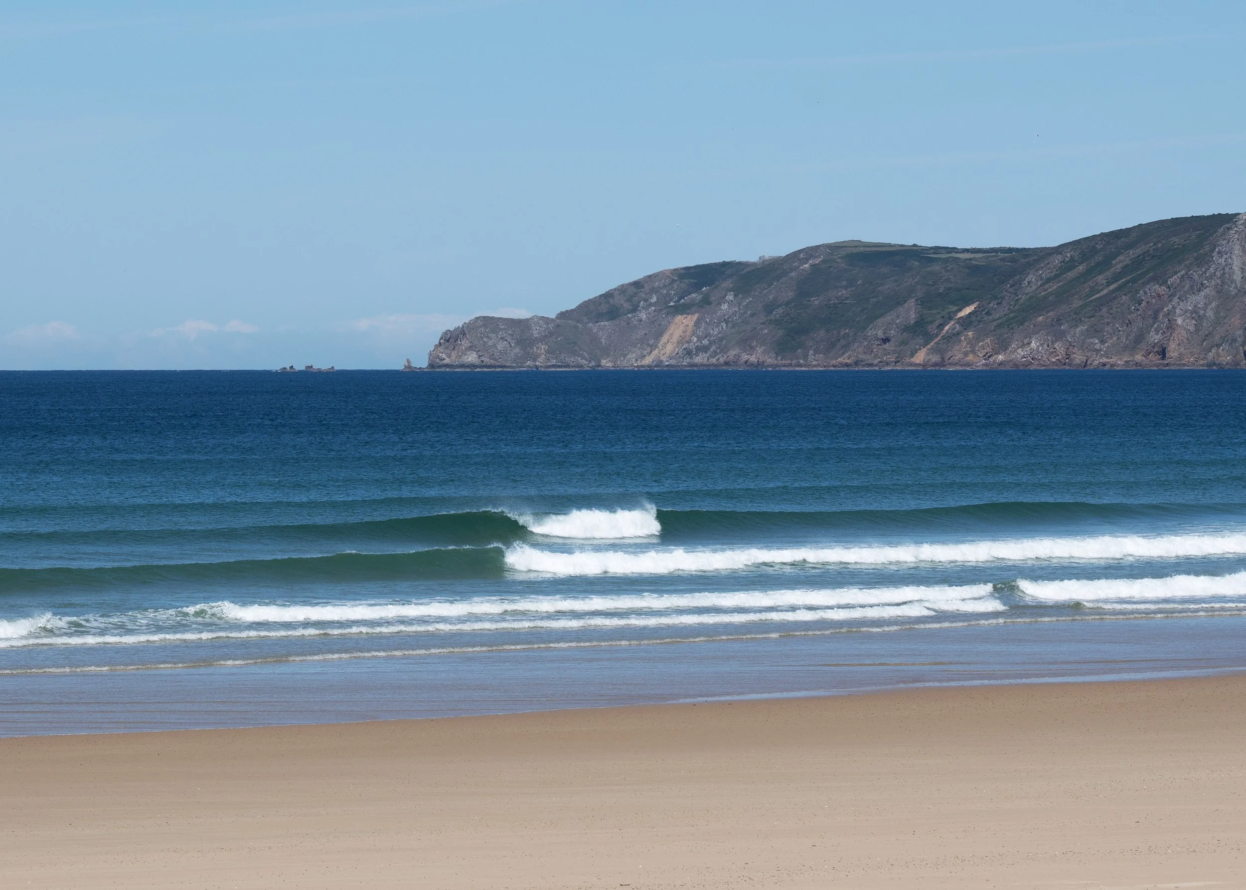 Une plage avec du sable, des vagues qui arrivent et une mer calme, avec des falaises en arrière-plan sous un ciel clair.