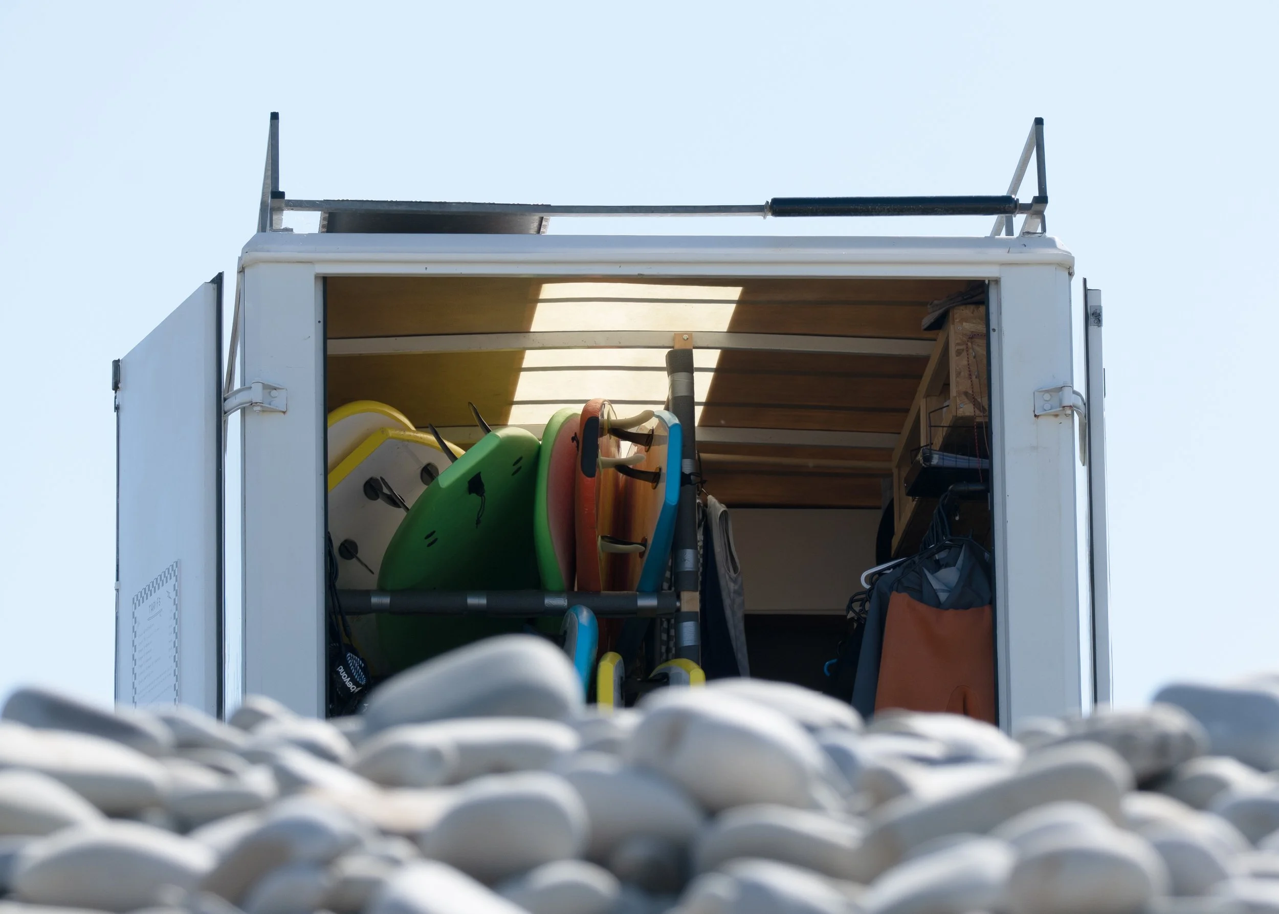 Le camion de la Vauville Surf School garée à sa place sur le Parking de la devise lors d'une belle journée ensoleillée, avec des galets au premier plan.