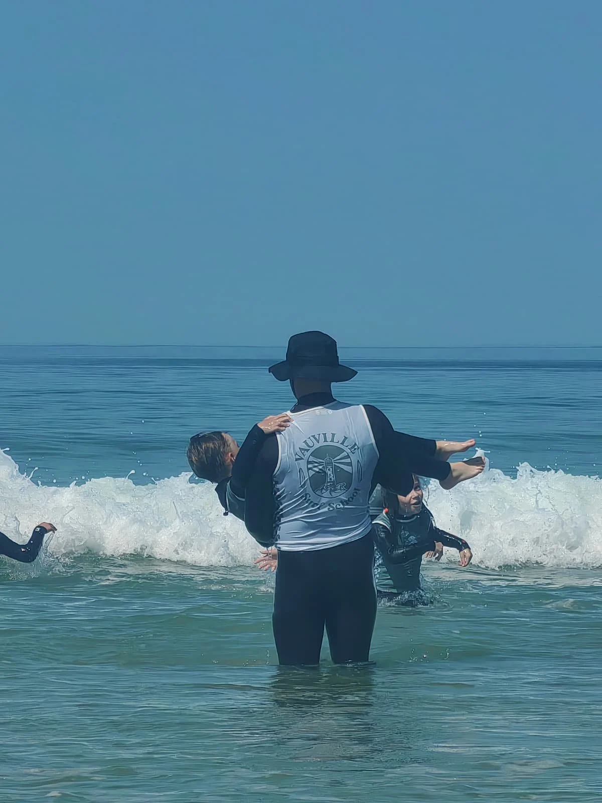 Le moniteur de surf portant un enfant au dessus des vagues, avec d'autres enfants nageant ou jouant dans la mer.