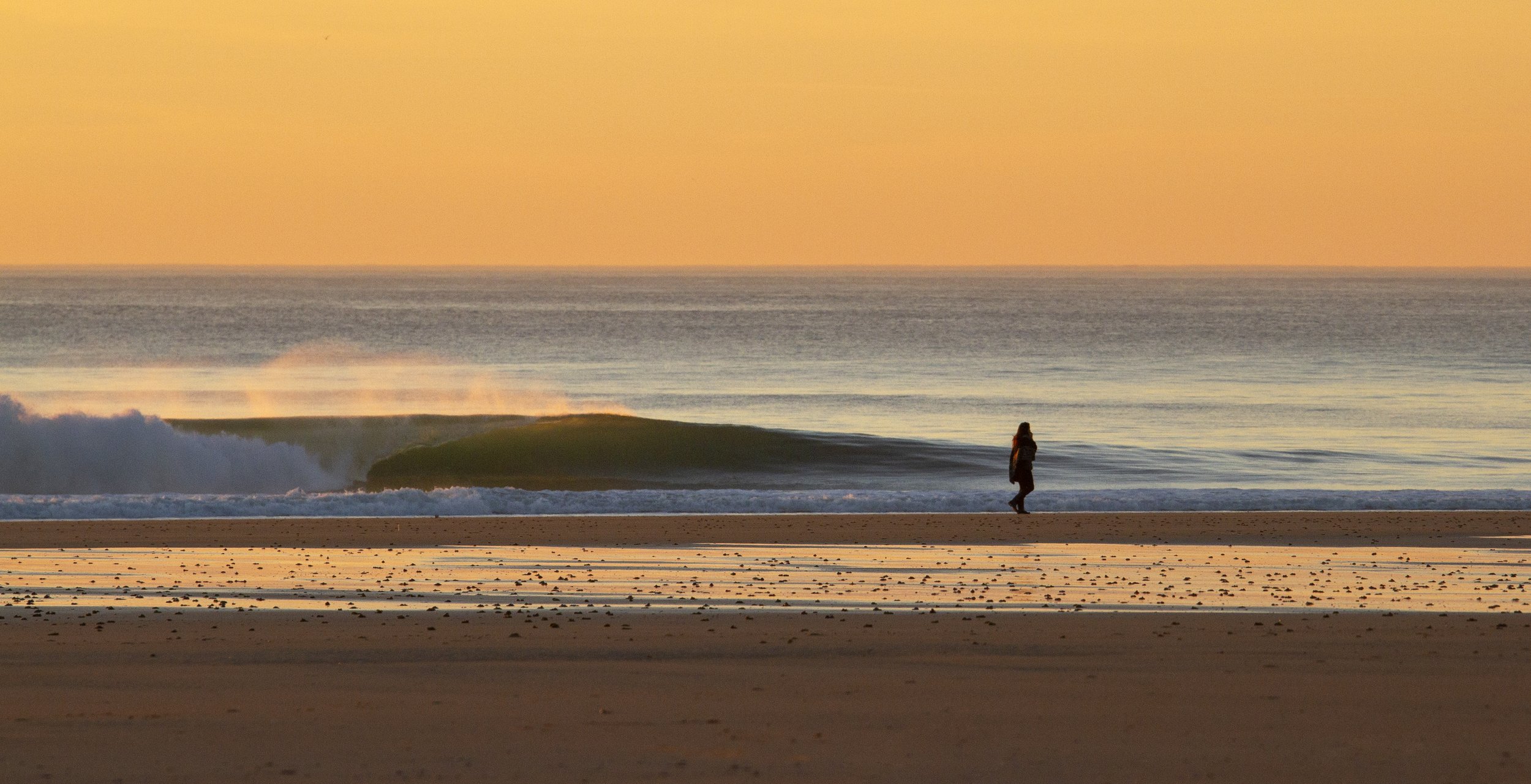 Une personne marche sur une plage au coucher du soleil, avec des vagues qui se brisent et un ciel orange.