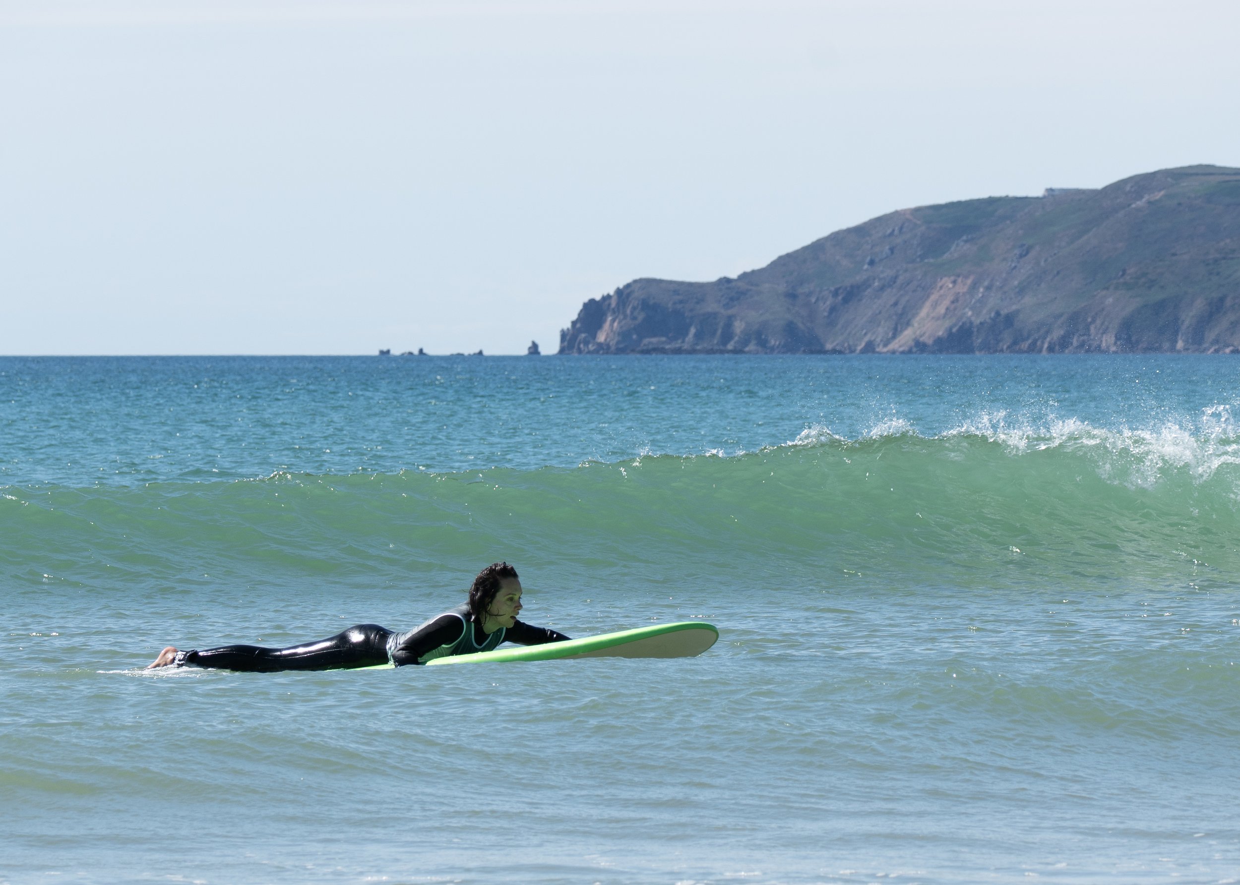 Une femme en combinaison de surf rame sur une planche de surf dans l'océan, avec une côte rocheuse en arrière-plan.