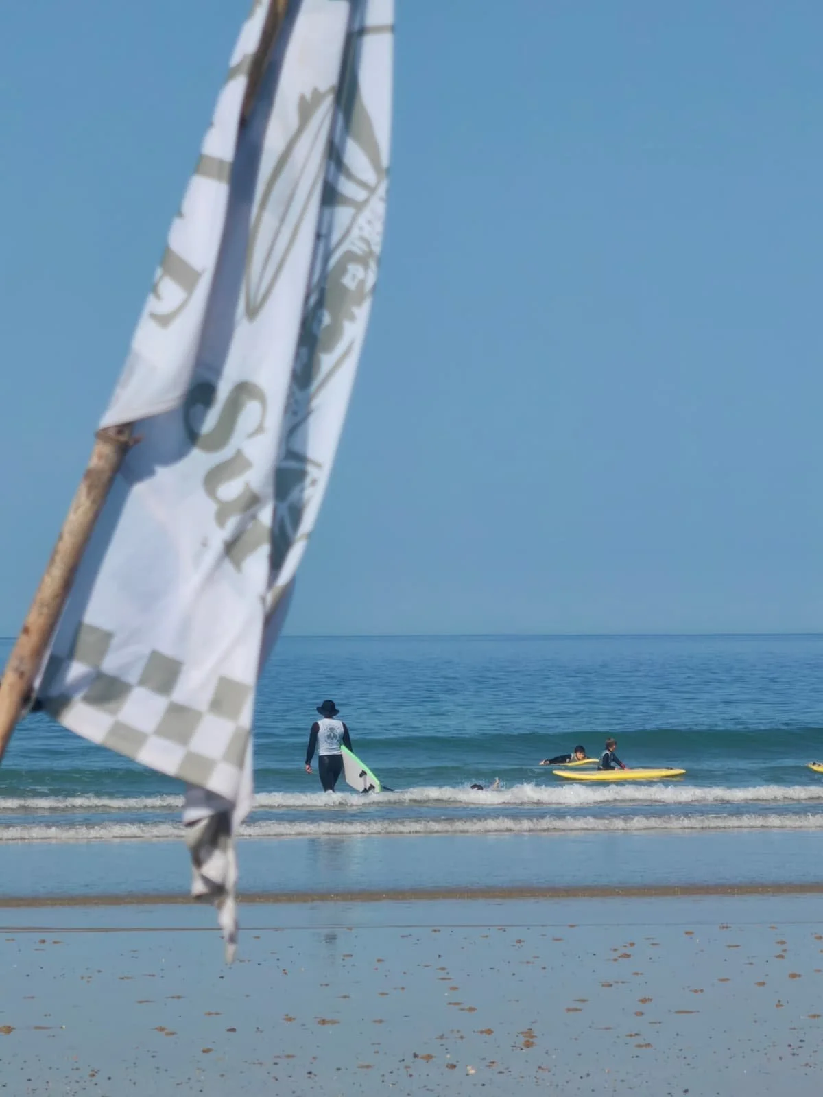 Une plage avec des personnes pratiquant le surf dans l'océan, un drapeau blanc  au premier plan, et un ciel clair.
