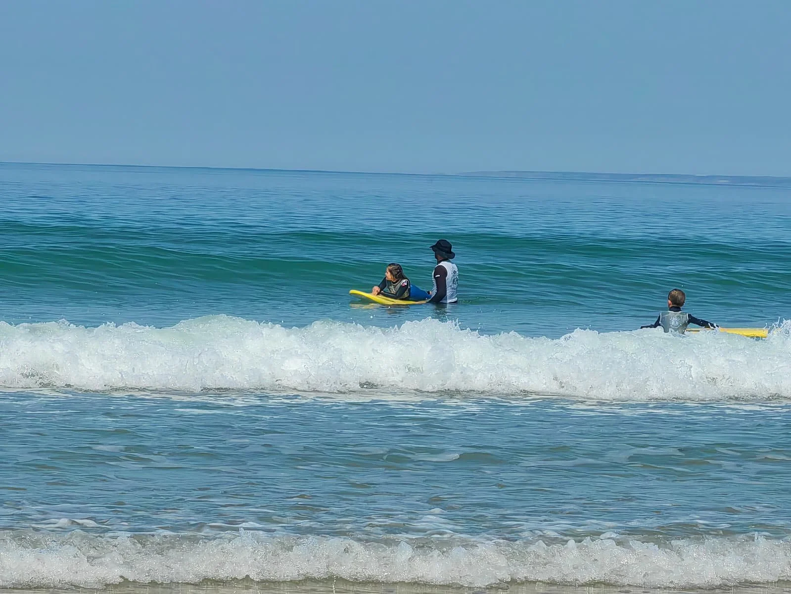 Trois personnes apprennent à faire du surf dans l'océan, deux d'entre elles étant debout sur leur planche et une étant assistée par un moniteur.
