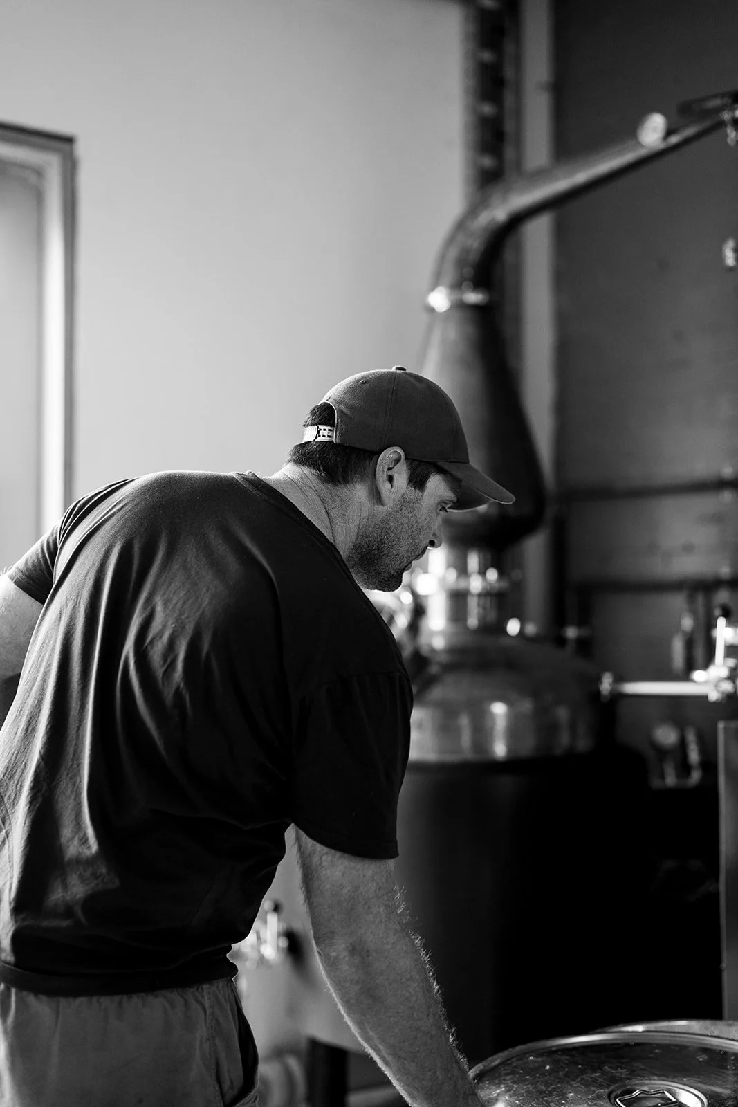 A man wearing a baseball cap and black t-shirt working in a kitchen with industrial equipment in black and white.