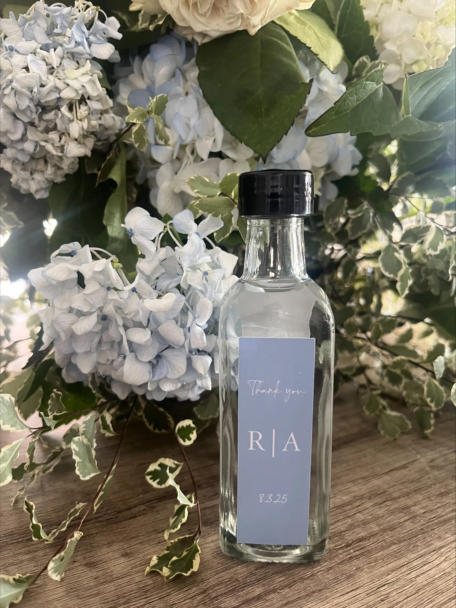 Empty glass bottle with black cap and a personalized label reading 'Thank you R|A 8.3.25' placed on a wooden surface, with a background of white and light purple hydrangea flowers and green variegated leaves.