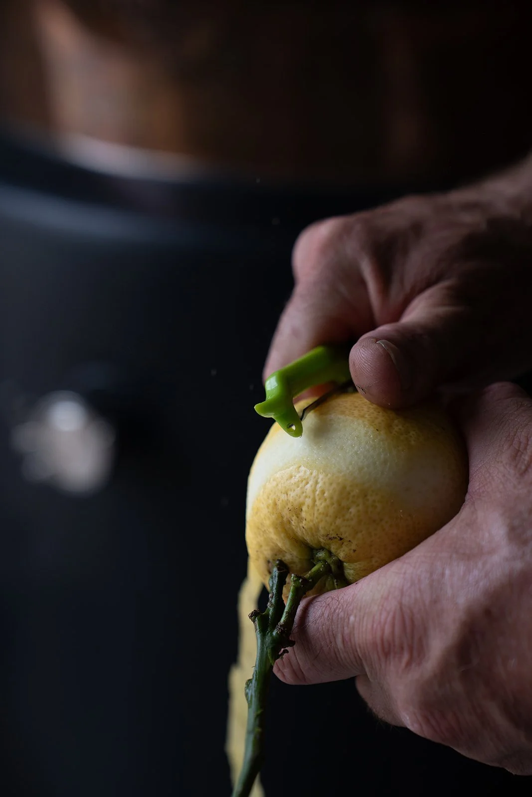 A person peeling a yellow lemon with a green vegetable peeler.