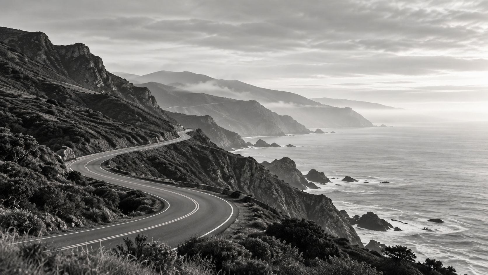 Black and white photo of a winding coastal road along rugged cliffs with the ocean on the right and mountains in the distance.