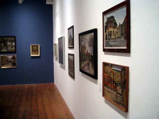 Gallery wall with framed paintings and photographs inside an art gallery with hardwood flooring.