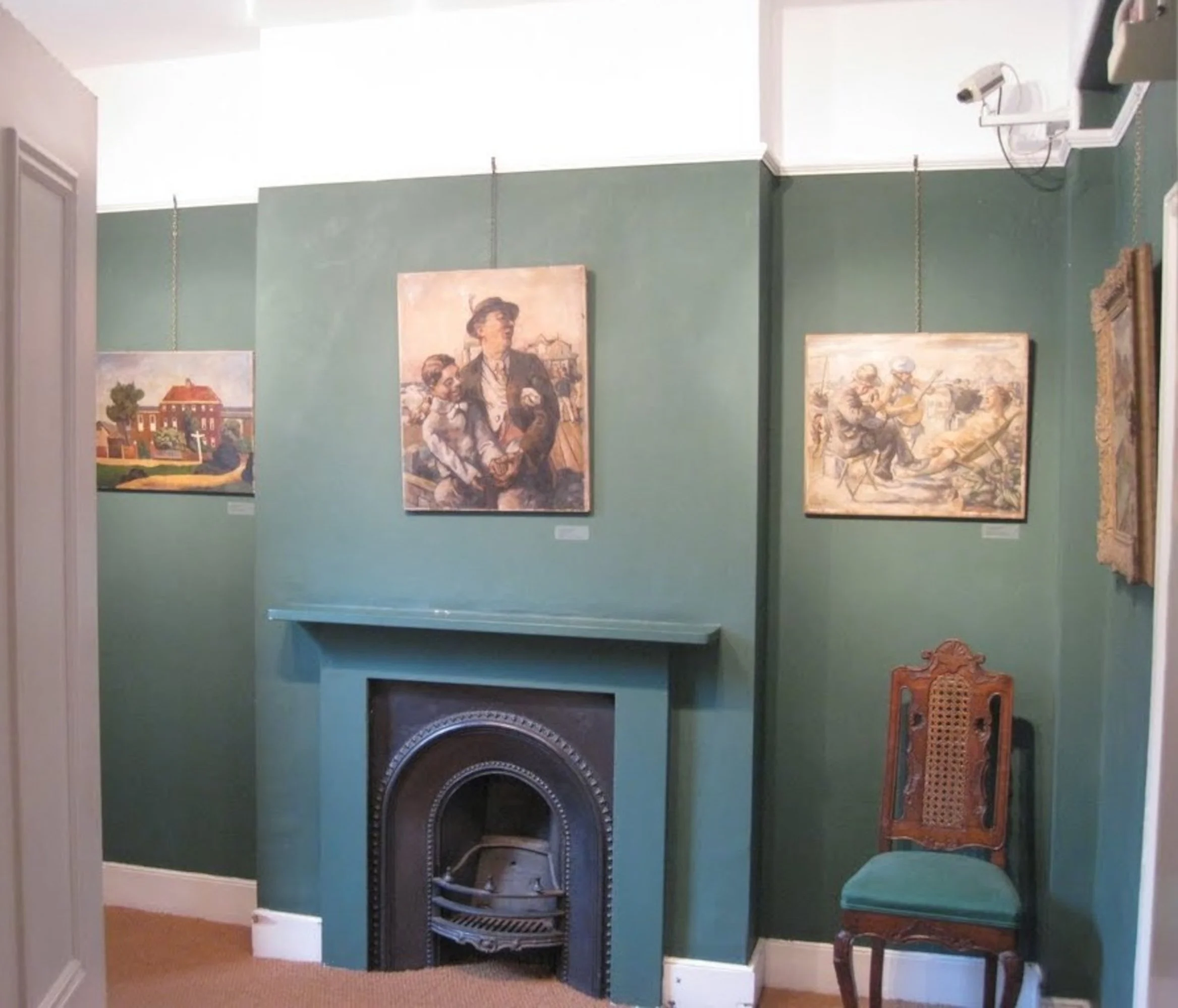 A room with green walls featuring three framed paintings, a small decorative fireplace, and a wooden chair with a green upholstered seat.