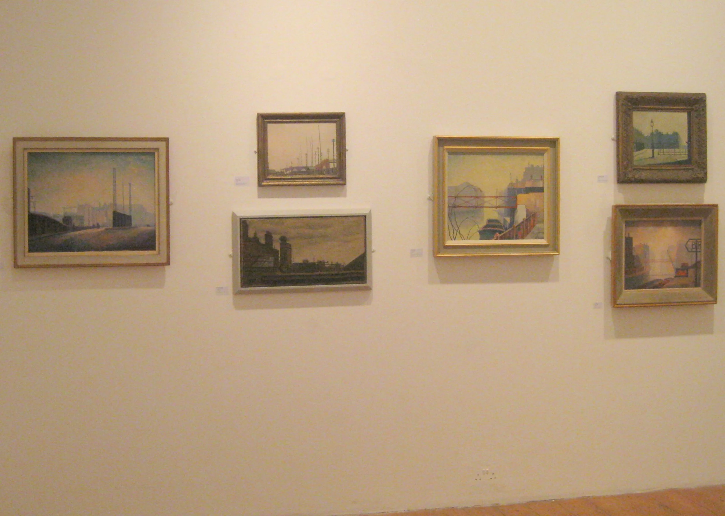 A white gallery wall with seven framed landscape paintings depicting industrial and harbor scenes, arranged in a mostly horizontal layout.