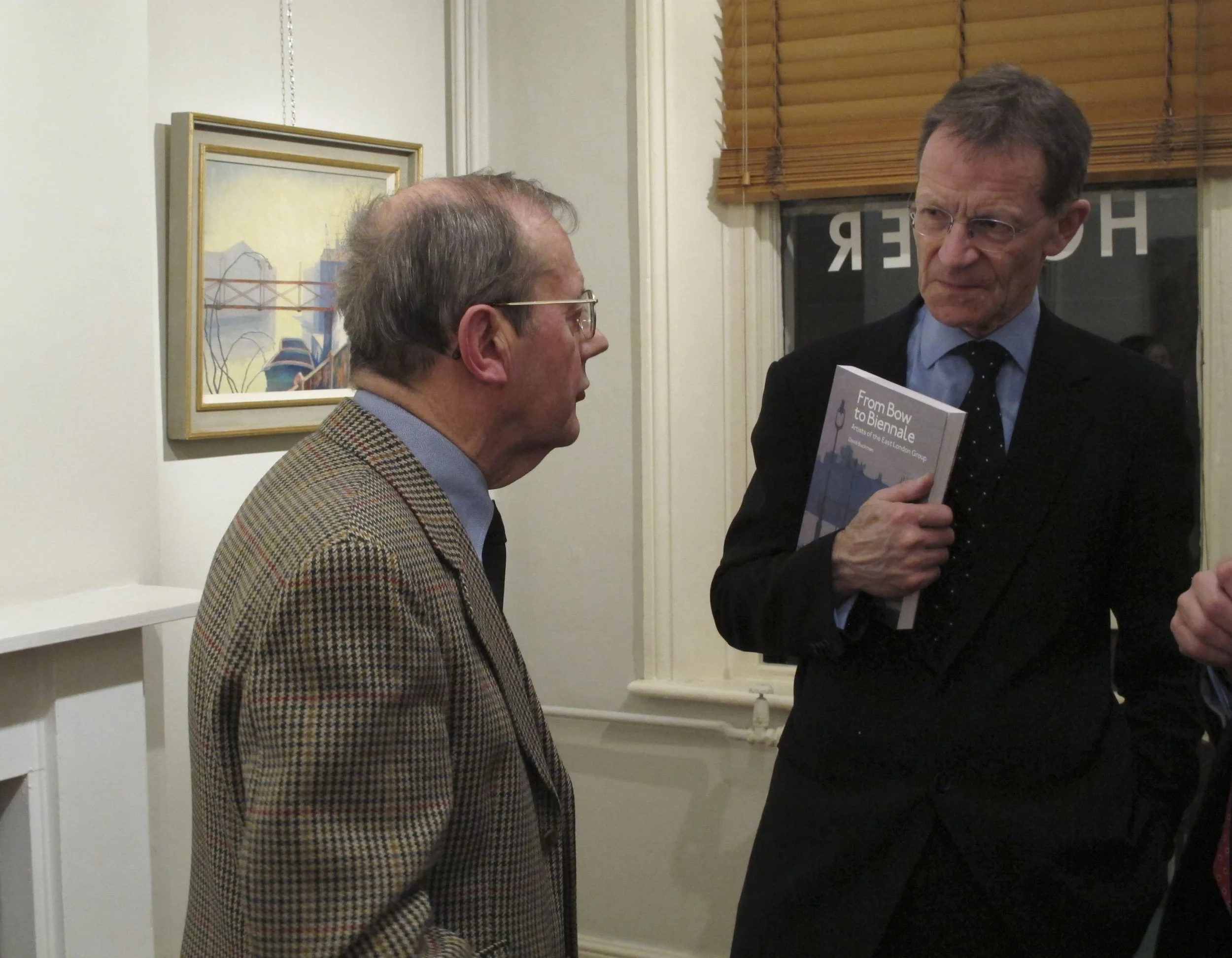 Two men in conversation indoors, one holding a book titled "From Bow to Biennale" in front of a window with blinds, and a painting on the wall.