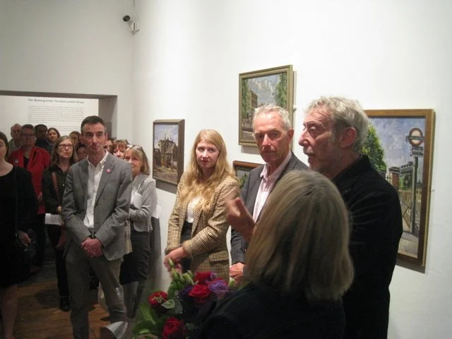 A woman with gray hair speaking in front of an art gallery crowd, with paintings hanging on the white walls behind them.